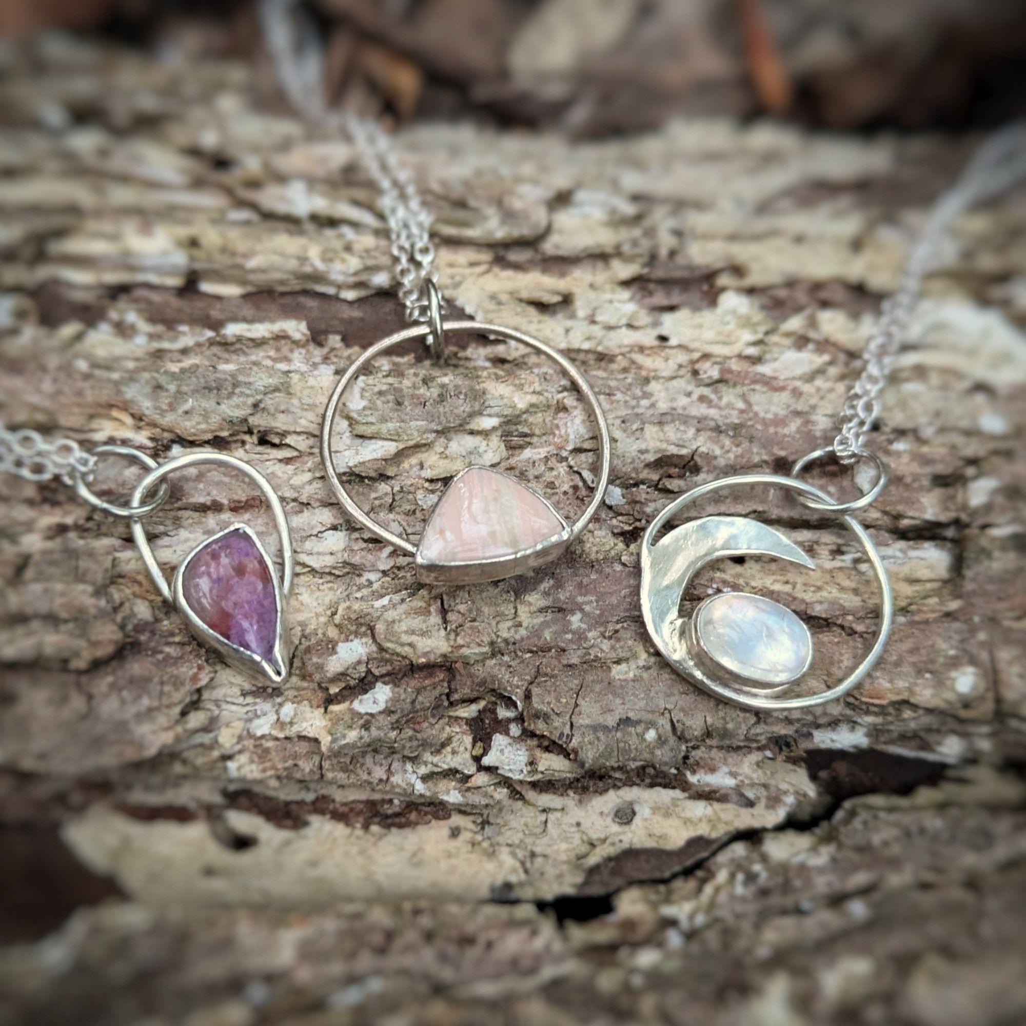 Three silver necklaces with large round bails hold small stones in different shapes: an asymmetrical teardrop shaped purple sugilite, a triangular pastel bacon looking stone, and an oval moonstone set in the crook of a crescent moon.