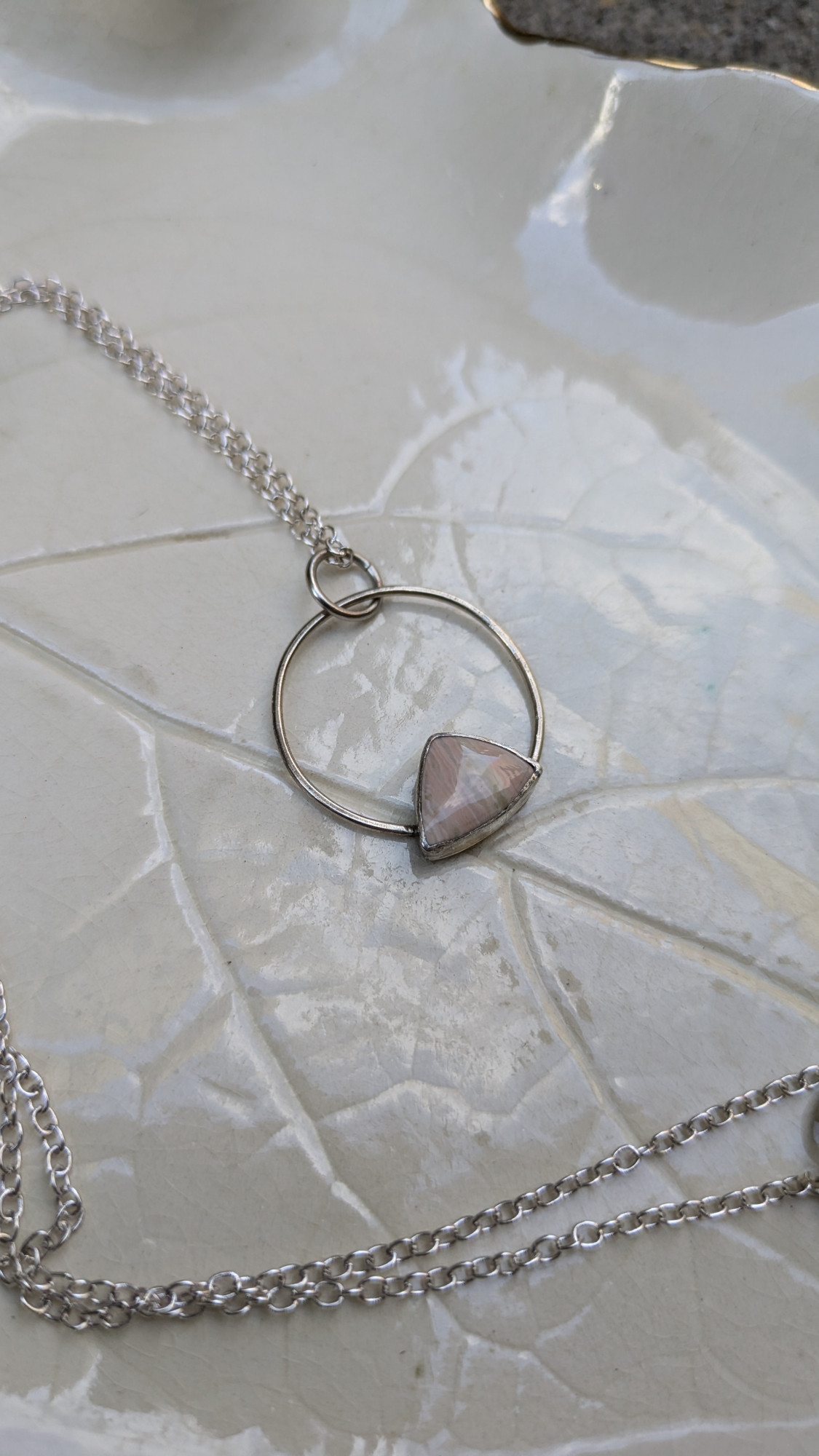 A silver necklace has a triangle shaped pink and cream striped stone set in a silver bezel on a large open circle bail.