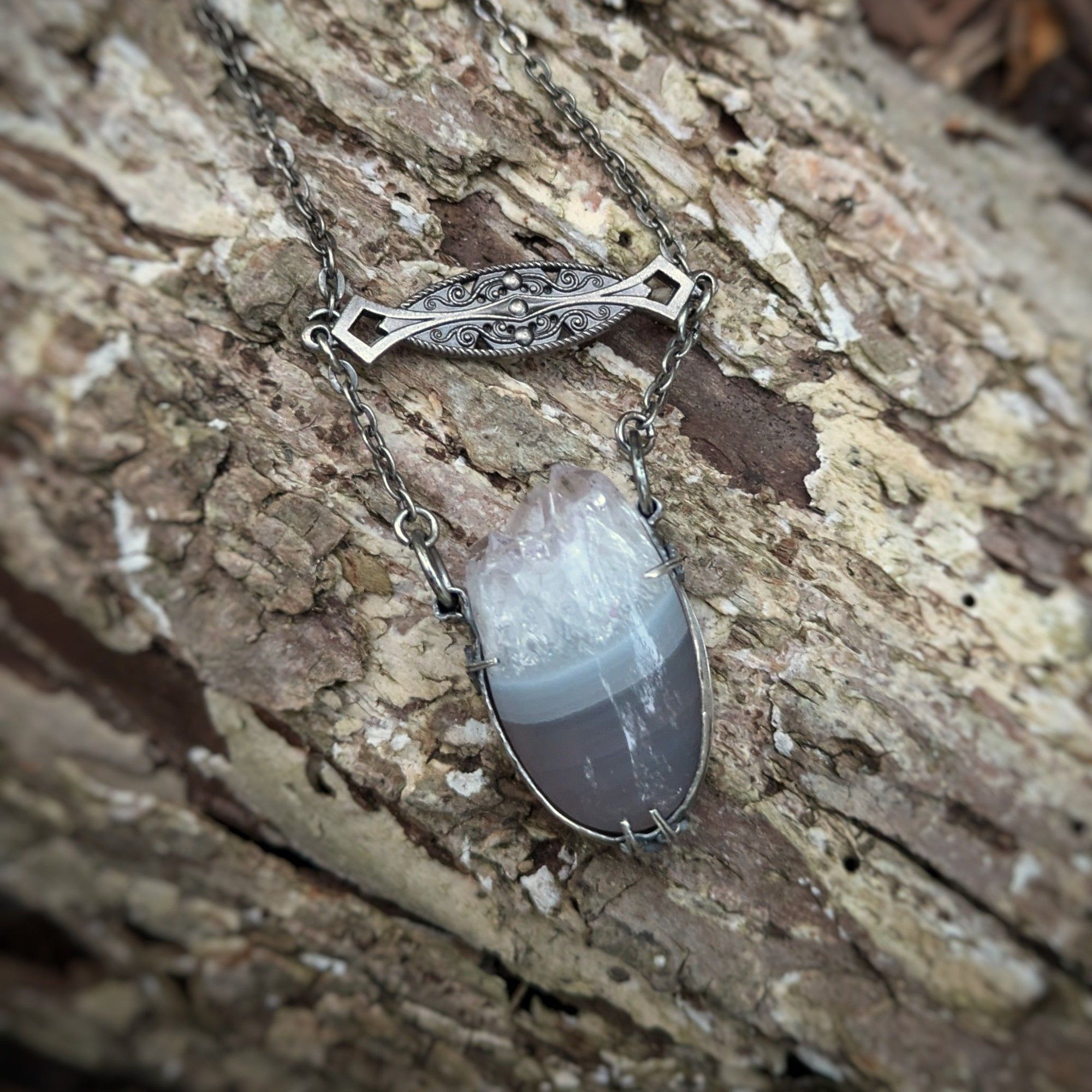 A blue banded asymmetrical agate with a live crystal edge is held in a silver frame with Gothic spikes holding it in place under a filigree connector and flat circular chain.