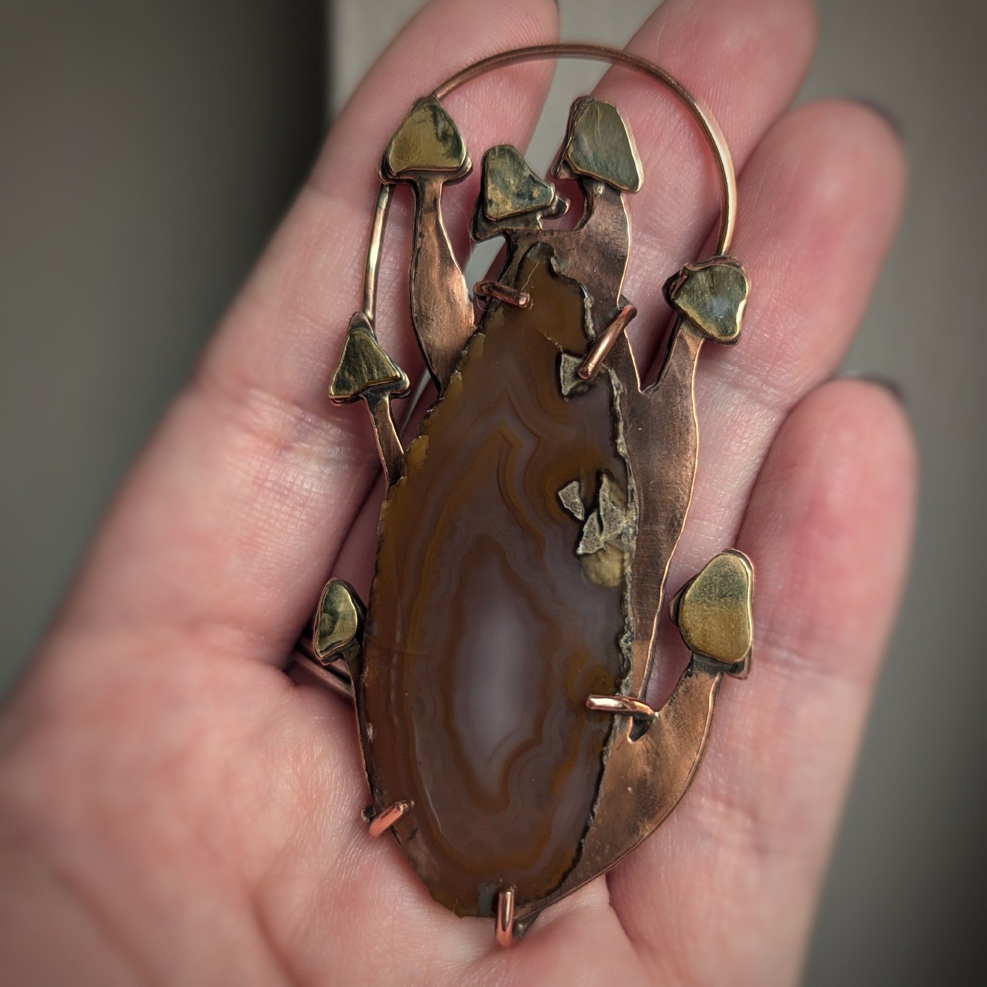 A copper pendant with brass capped mushrooms seeming to grow out of an agate slice which looks like a log.