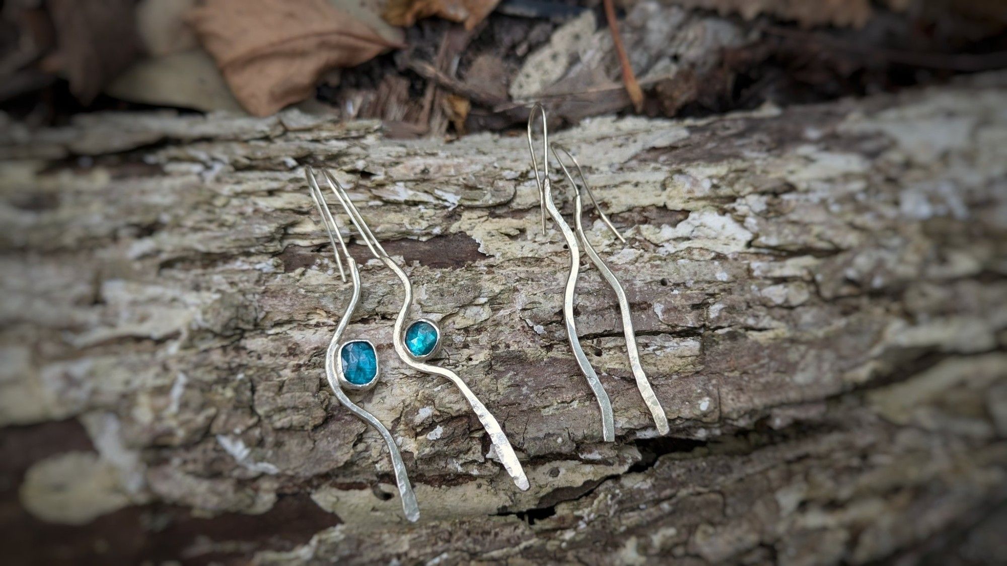 Two pairs of wavy silver earrings with a hammered texture, one solid silver and the other set with teal faceted kyanite stones.