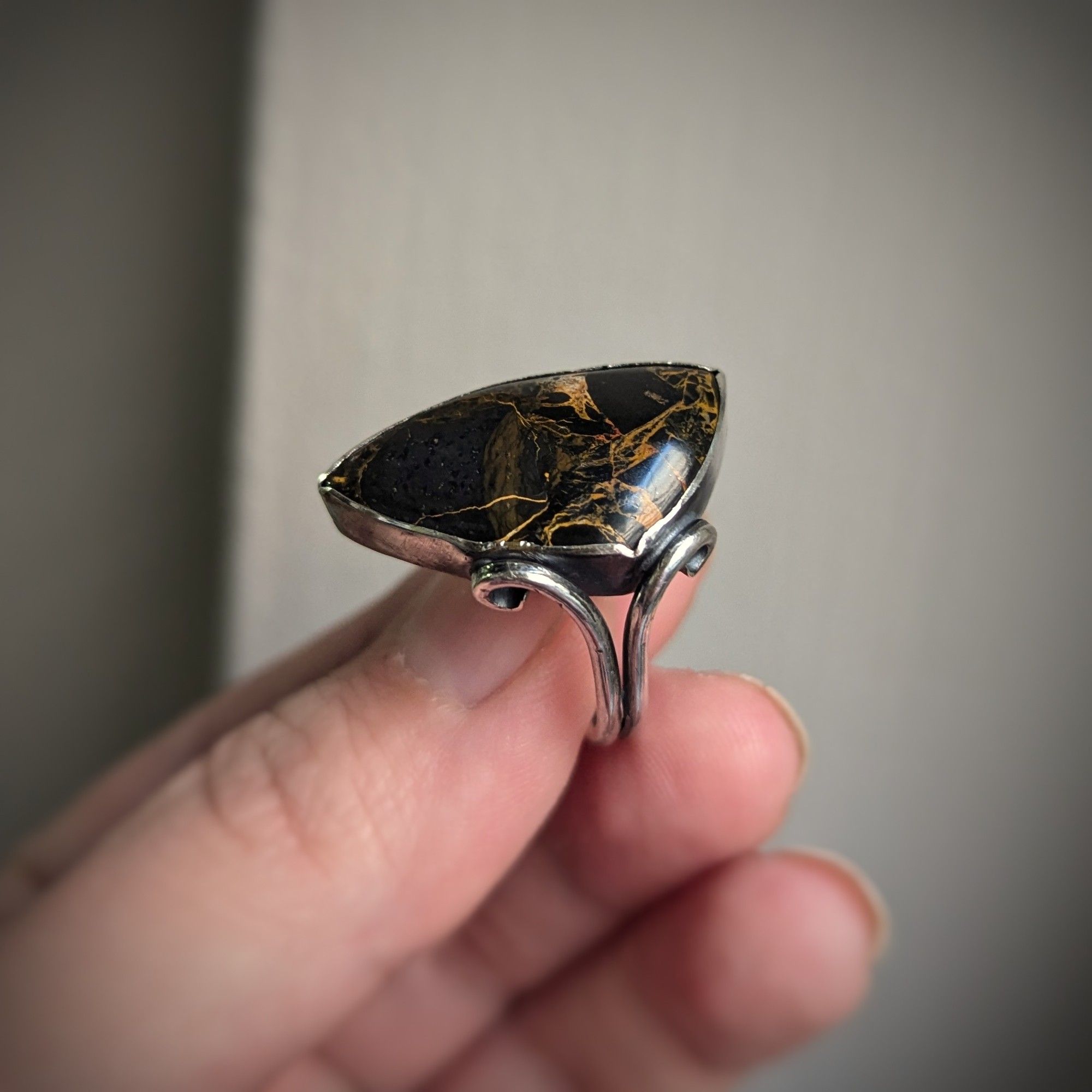 A black triangular stone with orange swirls is set in silver with the two wire ring shank extending up the side to form a swirling frame on one corner.