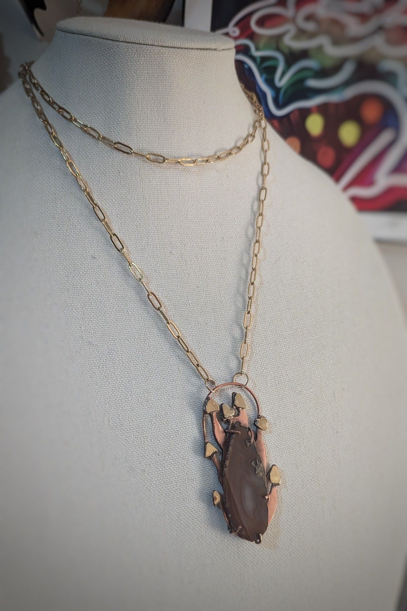 A copper and brass necklace with a large mushroom pendant holds a sliced agate in a prong setting, like a log sprouting a colony of mushrooms.