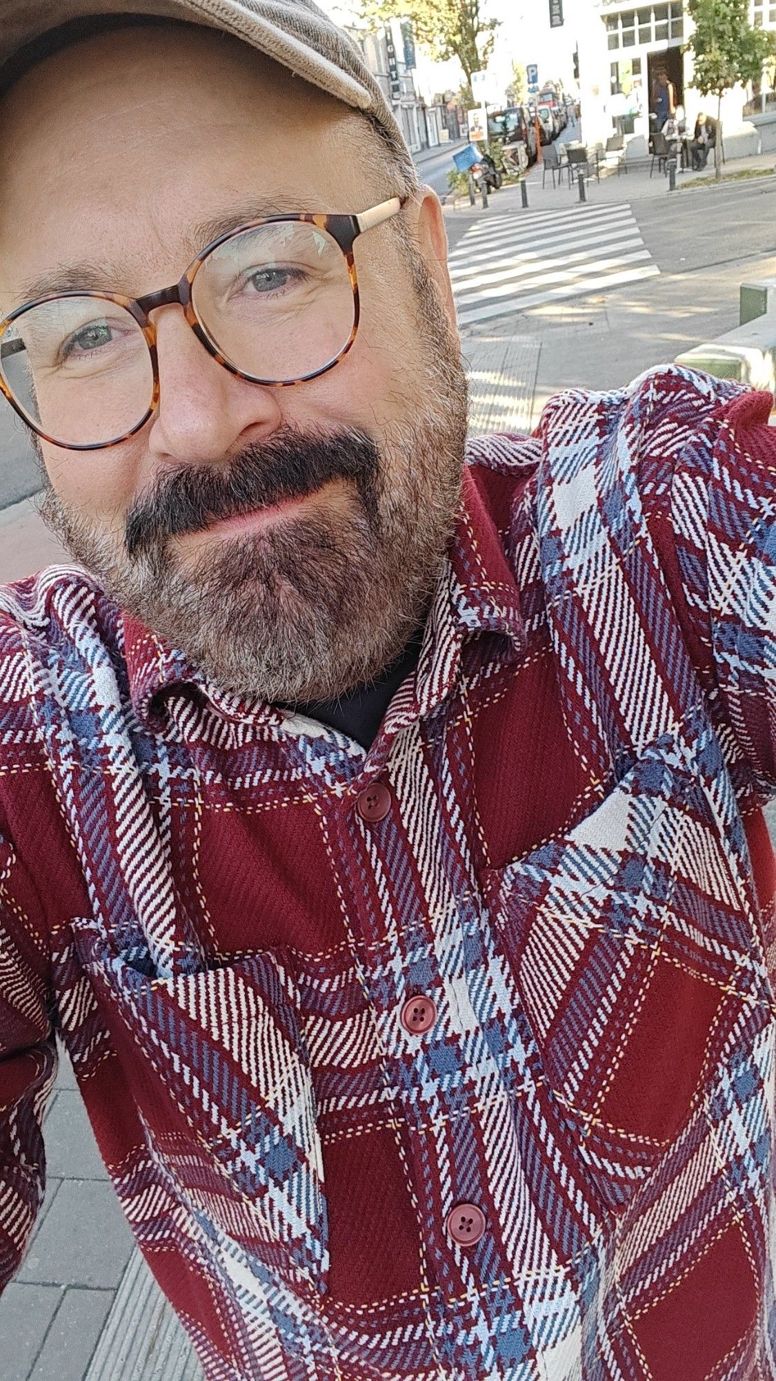 A selfie of me, a white bearded man, chubby, and wearing glasses, cap, and a blood-red plaid overshirt. I have the smile of a happy man.