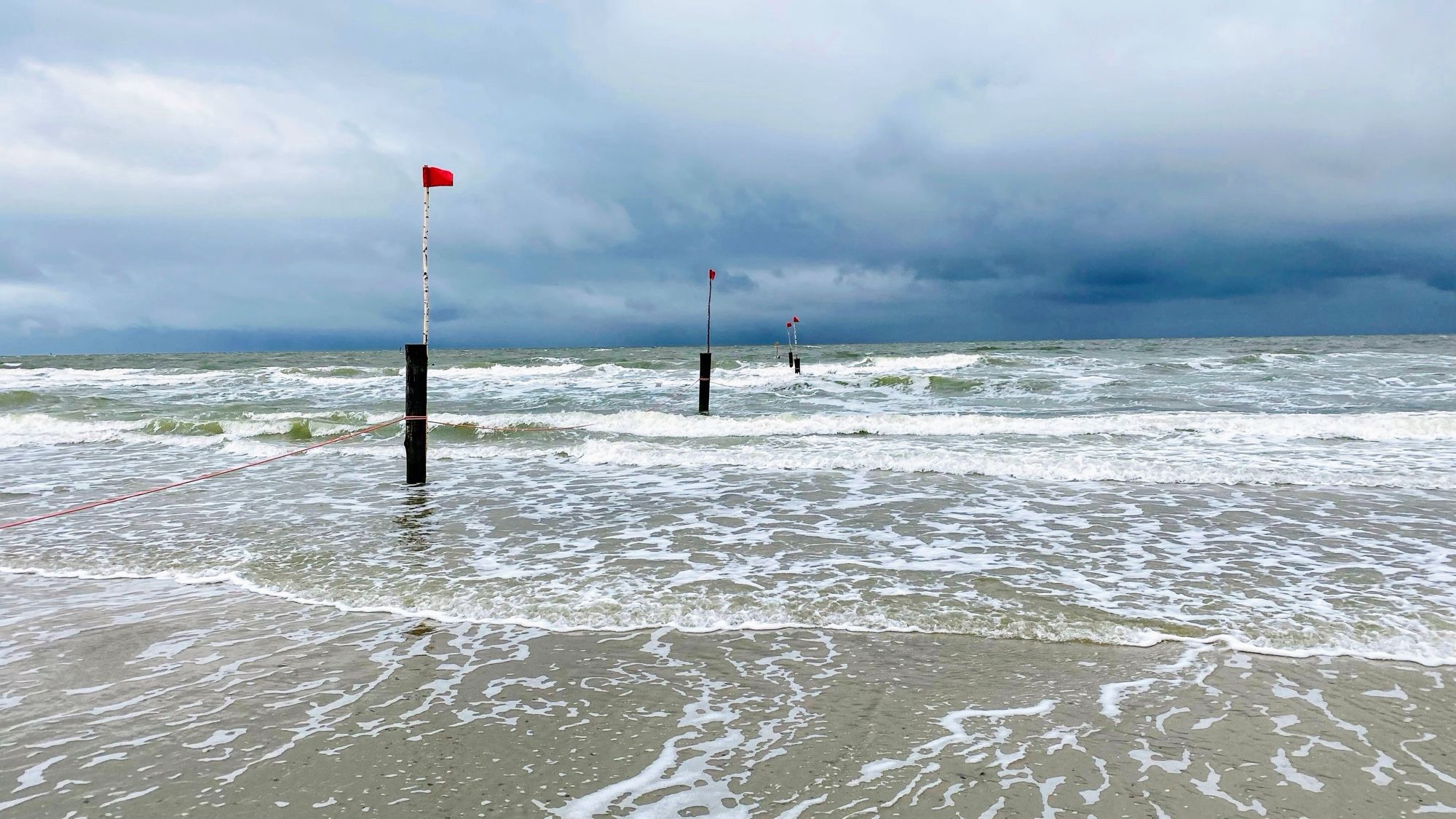 Rote Flagge auf einem Pfosten. Am Meer, Schaumkronen auf den Wellen. Dunkle Wolken.