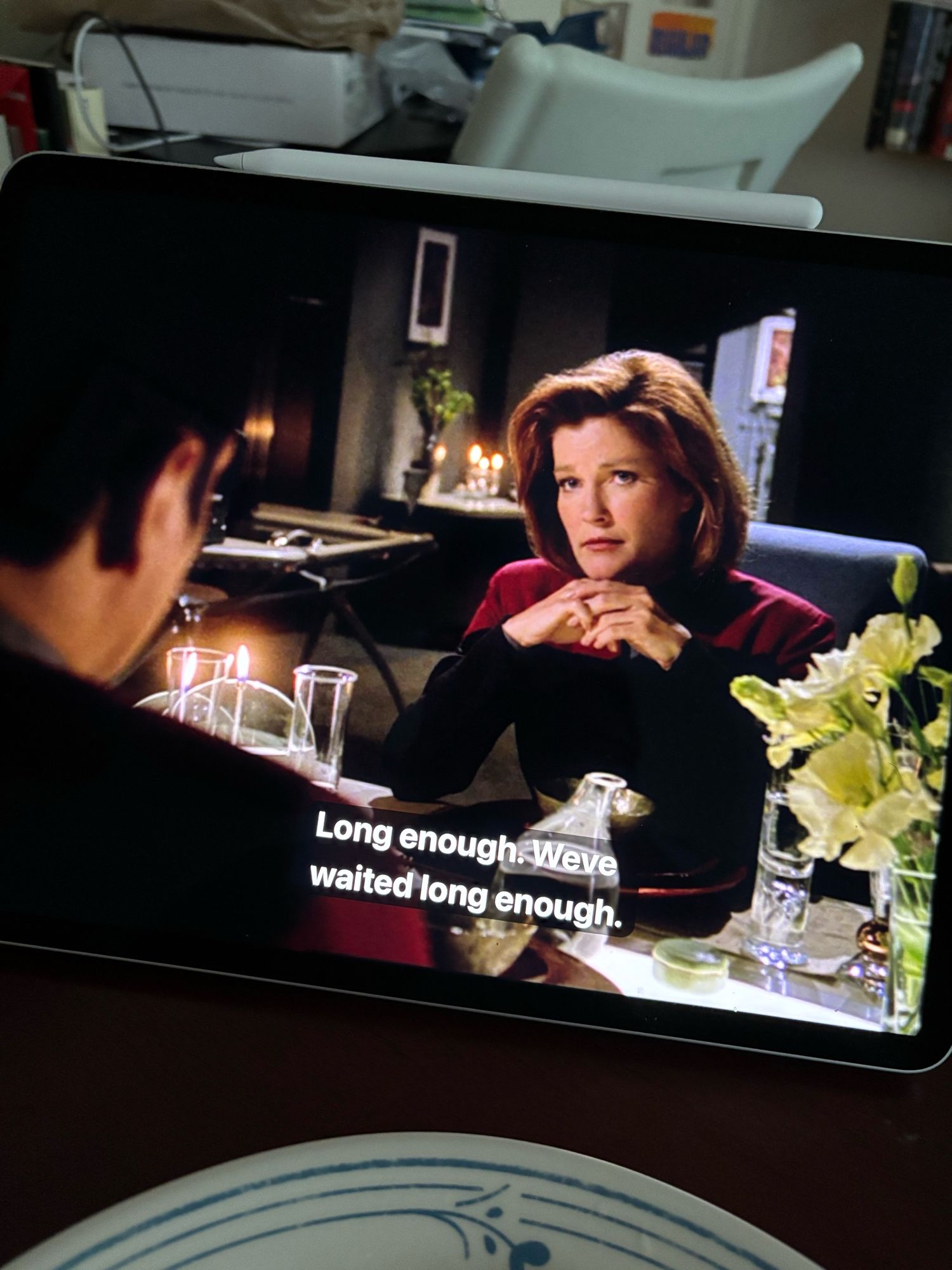 I took a picture of my iPad during a scene from the fifth season “Star Trek: Voyager” episode entitled “Timeless.” In this scene, Commander Chakotay is at foreground with his face turned away from the camera as he has dinner with Captain Janeway. The dinner table is decorated with green flowers of some sort, candles, and fine glass settings. In the background behind Captain Janeway, we can make out details of the interior her quarters, such as assorted furniture, assorted decor, and also more candles. At the dinner table, Captain Janeway sits with her hands folded together gazing thoughtfully at Chakotay. Two lines of closed captioning text read, “Long enough. We’ve waited long enough.”