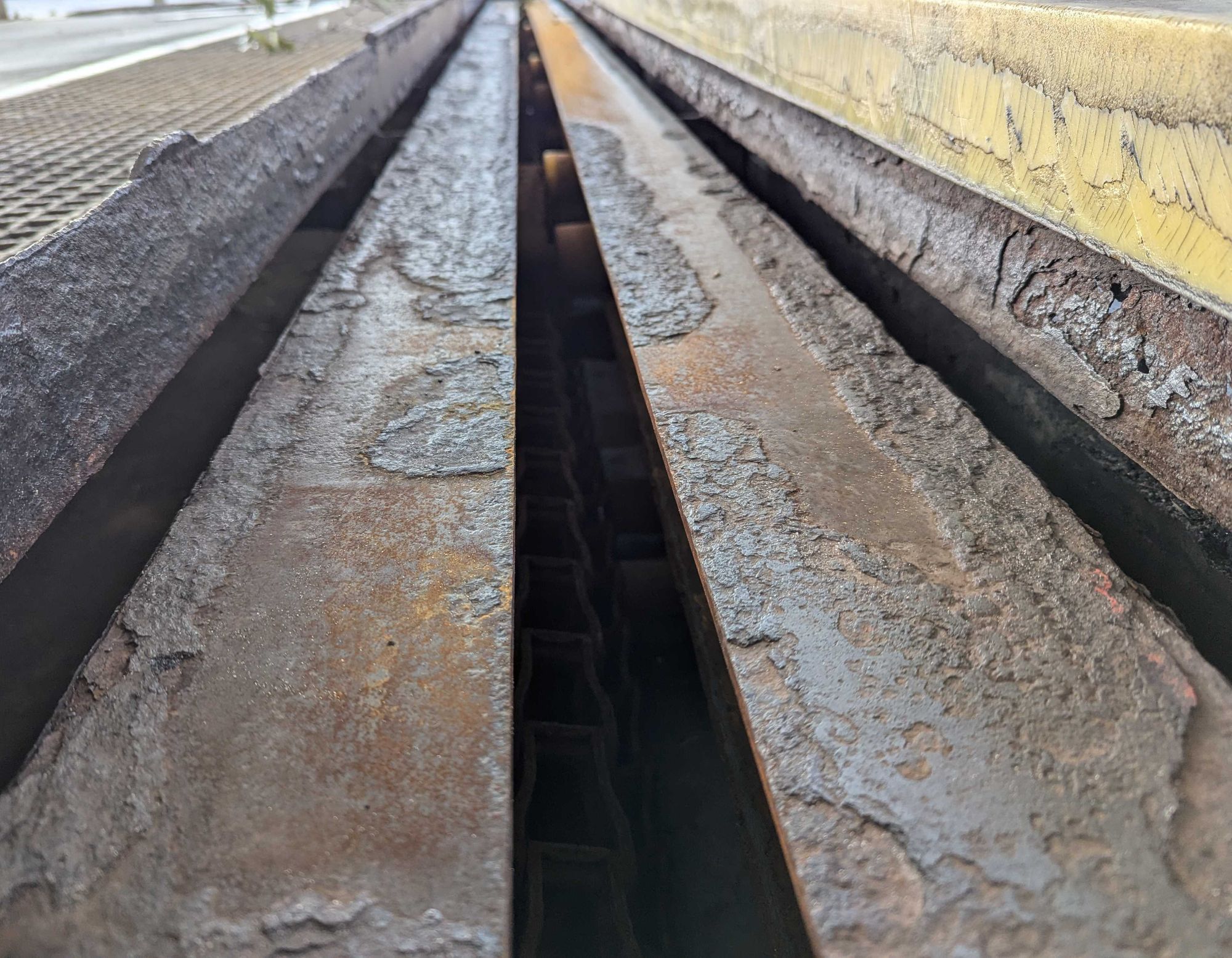 An old car wash tire guide, you can see the layers of rust on it, a drainage grill to the left with a large yellow curb to the guide to the right.