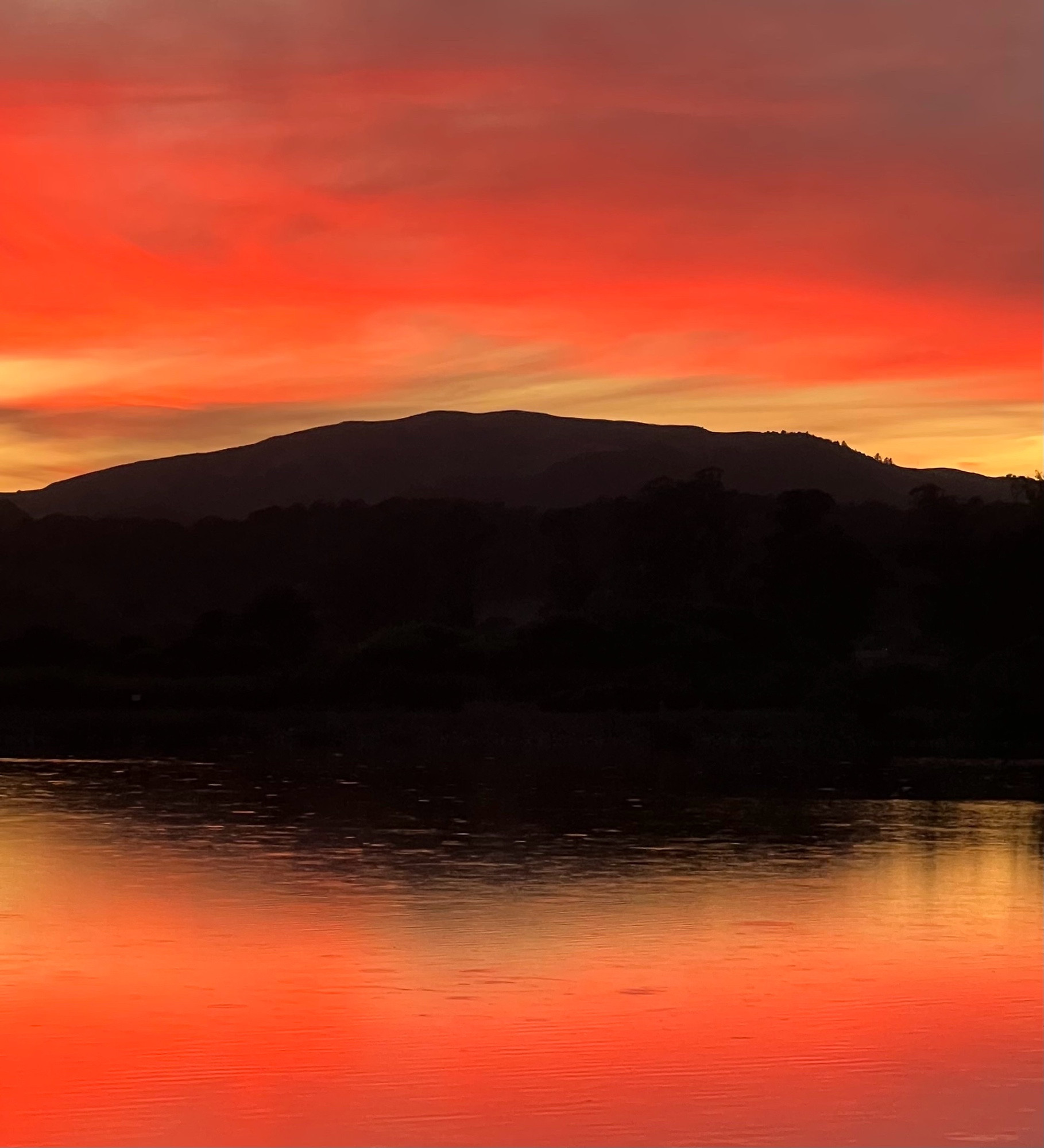 A stunning sunset with deep rose/orange hues of the sky reflected in the water, the two mirror images of color separated by a dark broad hill