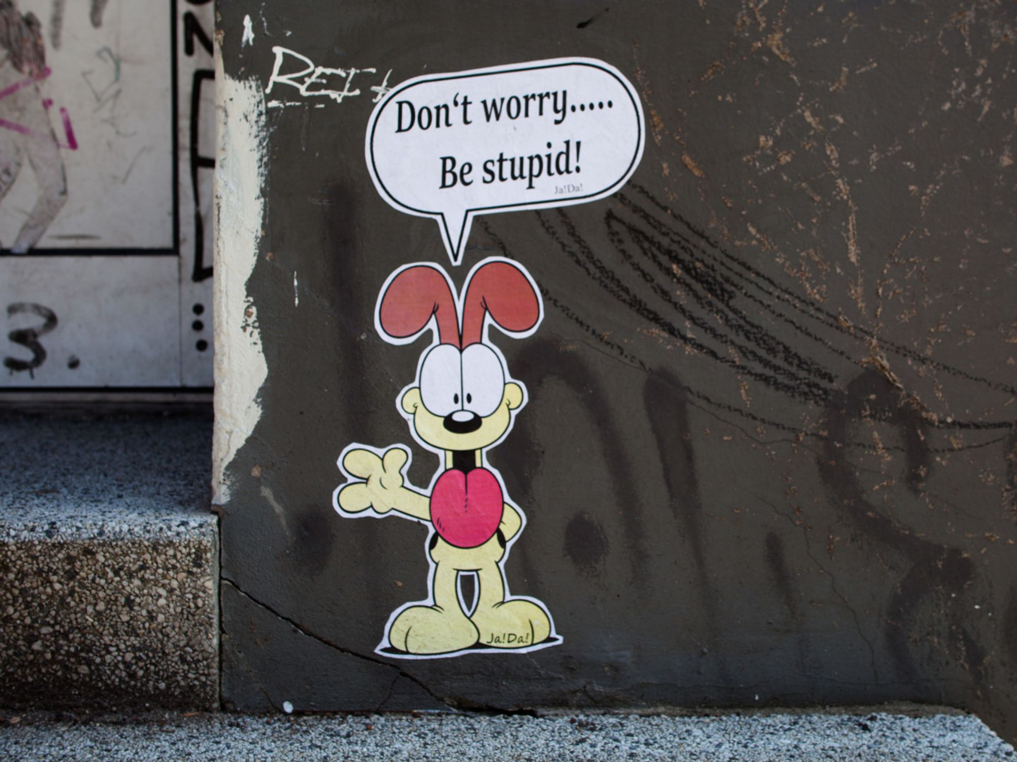 Sticker of Odie (from Garfield Comics), on an old grey wall, with a speech bubble "Don't worry… Be stupid!"