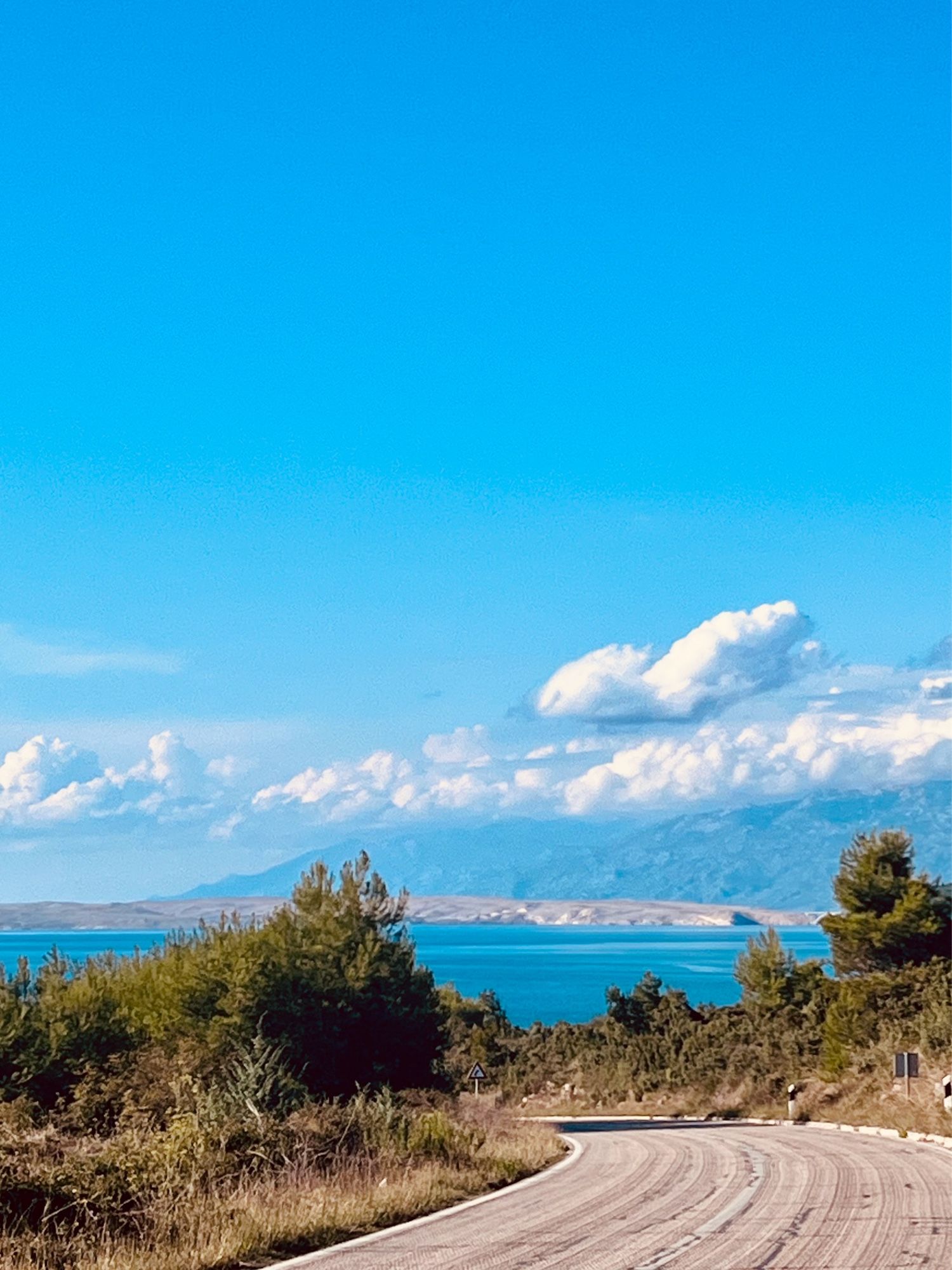 Eine Landstraße führt hinunter ans Meer, im Hintergrund die Insel Pag mit Brücke und das Velebit Gebirge, über dem sich einpaar Wolken halten. Ansonsten strahlendblauer Himmel im Spätsommer