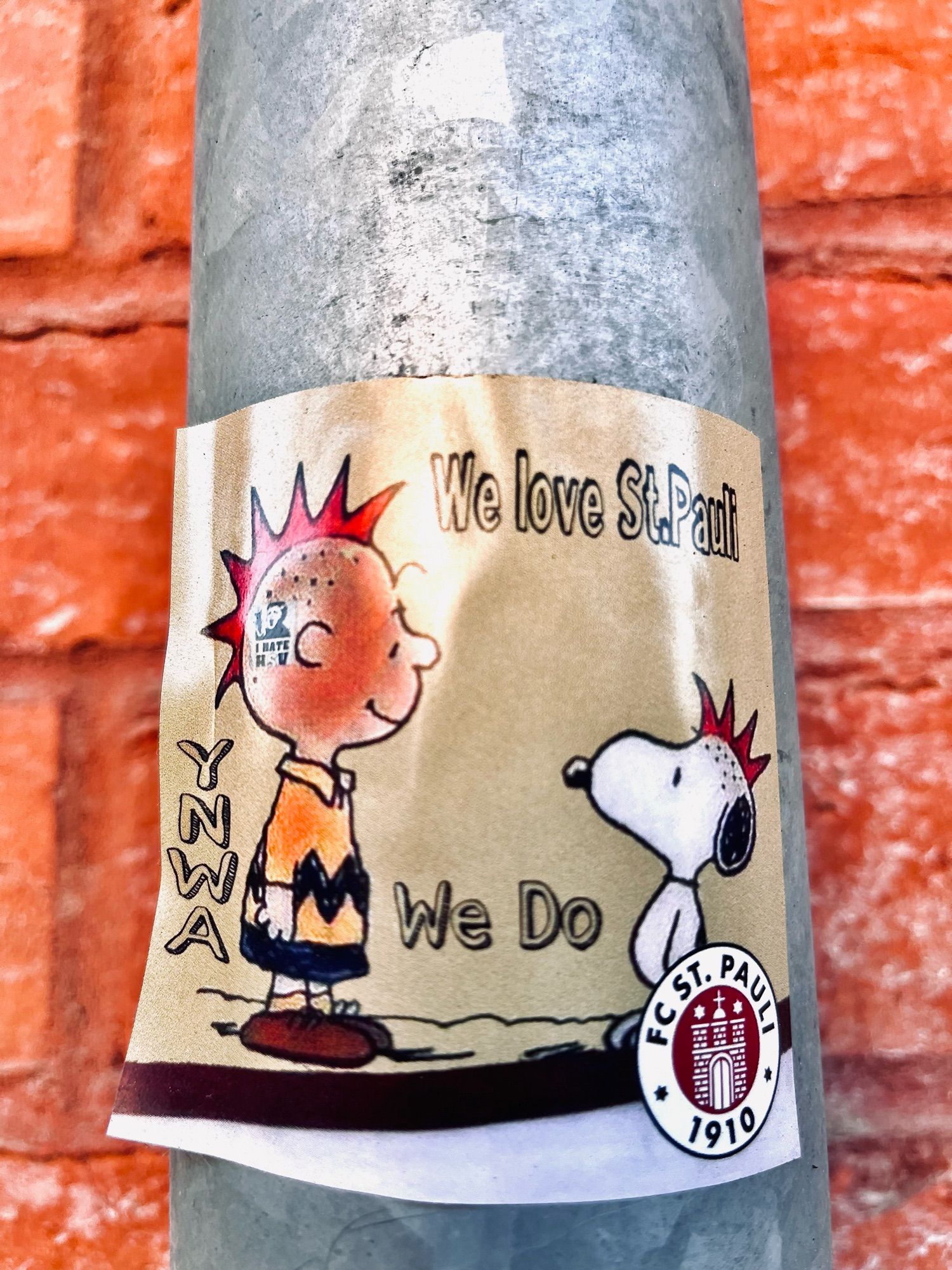 Sticker auf rot-verklinkerter Hauswand, bzw Regenrohr. Charlie Brown und Snoopy mit rotem Iro im, Aufschrift We love St.Pauli, You never walk alone in Initialien und Fußball Emblem FC.St.Pauli 1910