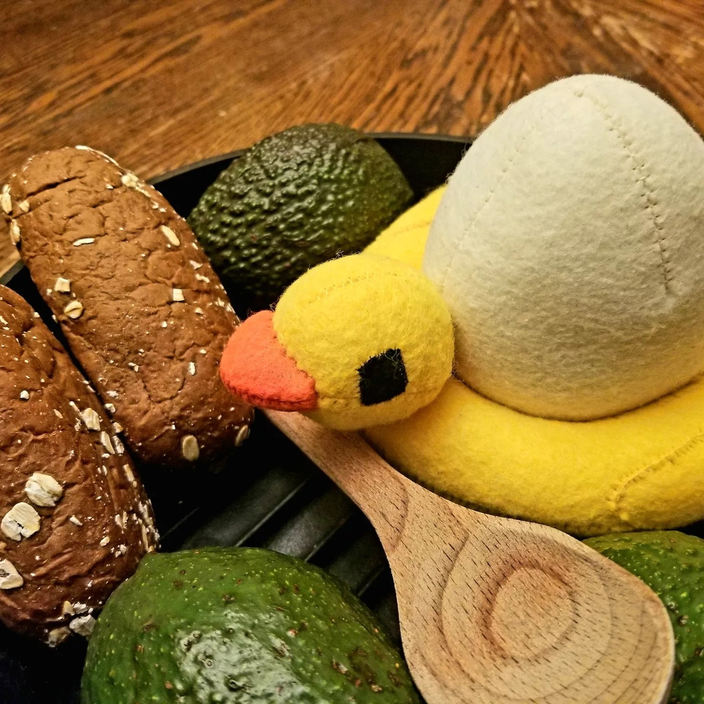 Similar subject, but at a different angle. Brown wooden table in the background of a black iron skillet containing three ripe avocados, two small loafs of bread, and a custom plush of QSMP egg character, Chayanne. Chayanne is a white egg with a bright yellow duck pool tube and little black stick legs. He sits with a wooden spoon in front of him.