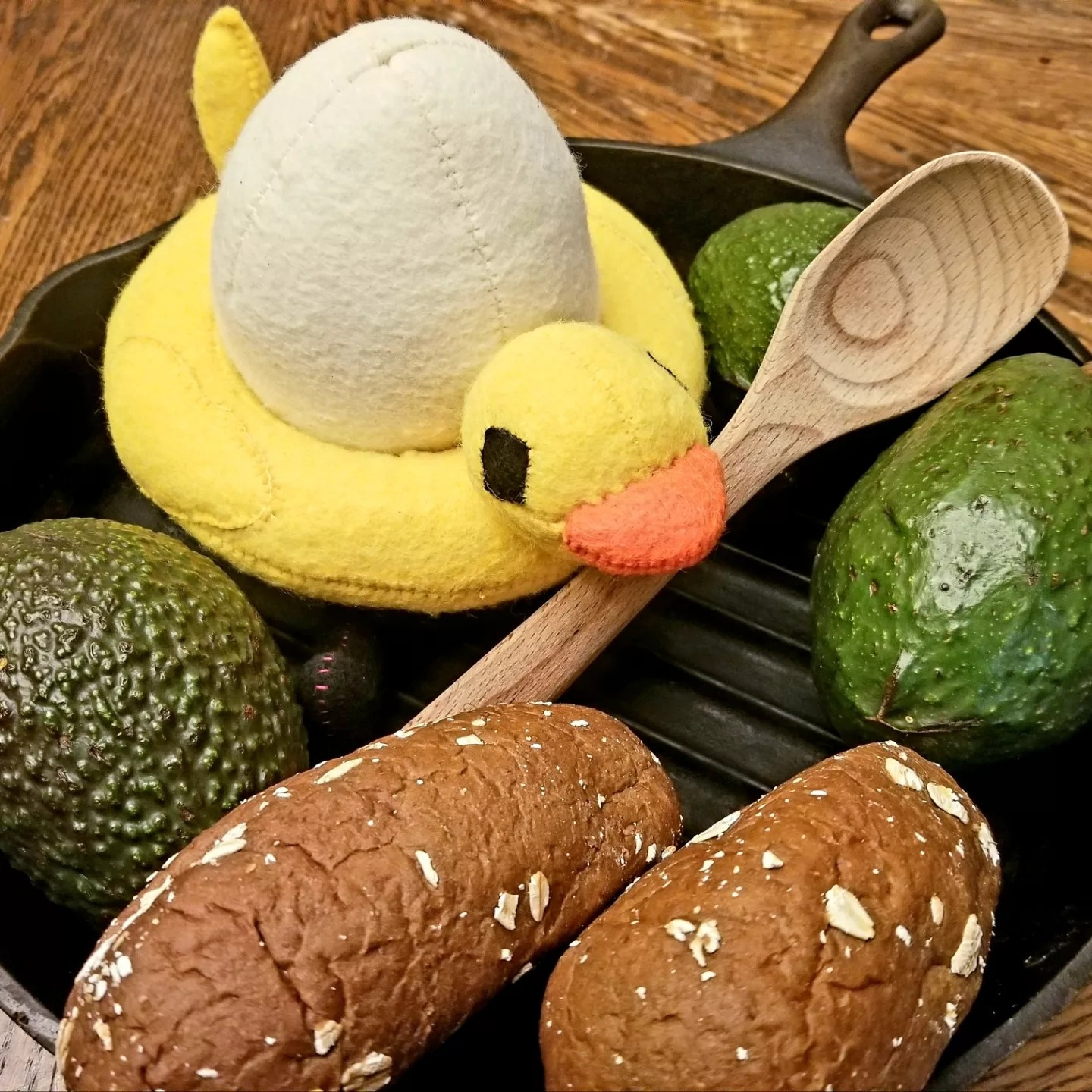 Brown wooden table in the background of a black iron skillet containing three ripe avocados, two small loafs of bread, and a custom plush of QSMP egg character, Chayanne. Chayanne is a white egg with a bright yellow duck pool tube and little black stick legs. He sits with a wooden spoon in front of him.