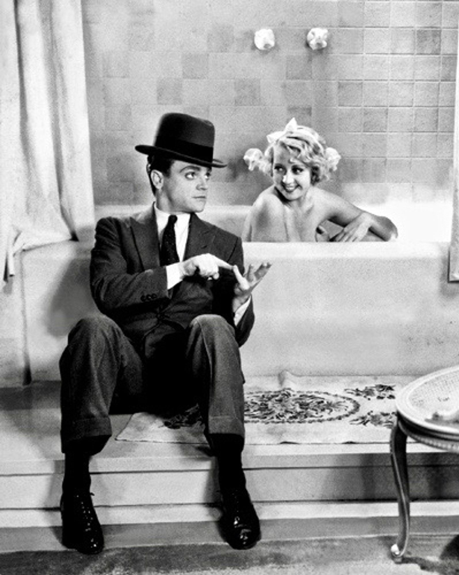 James Cagney sat on the floor in a suit and fedora next to a bath where Joan Blondell is washing herself