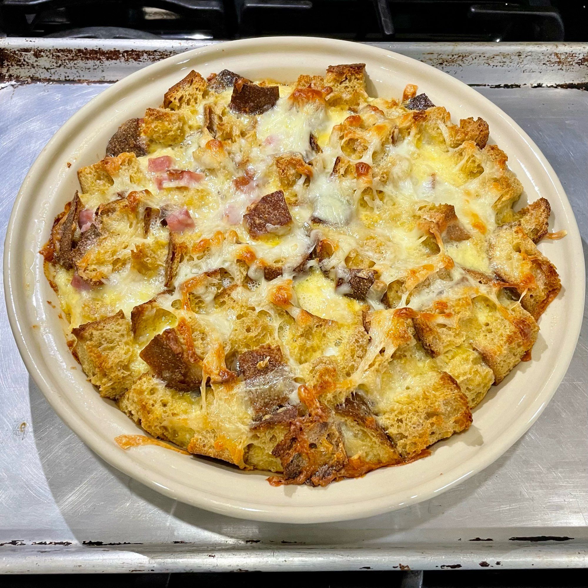 A cream colored pie pan filled with bread cubes, cheese, ham, and custard, baked until golden and crisp and gooey with melted cheese.