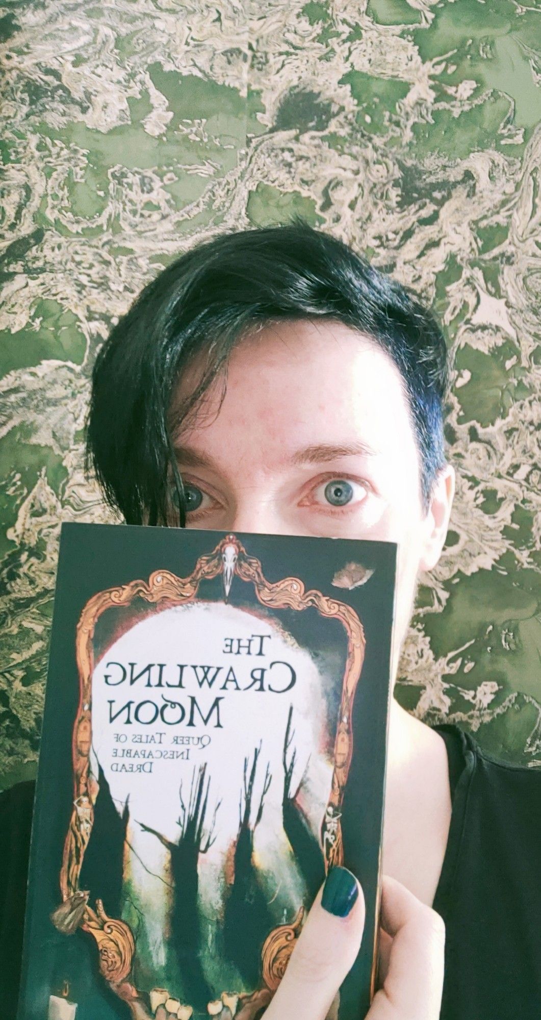 Someone peers over a book they're holding up, only eyes visible, looks a bit daunted. Title: The Crawling Moon, queer tales of inescapable dread.