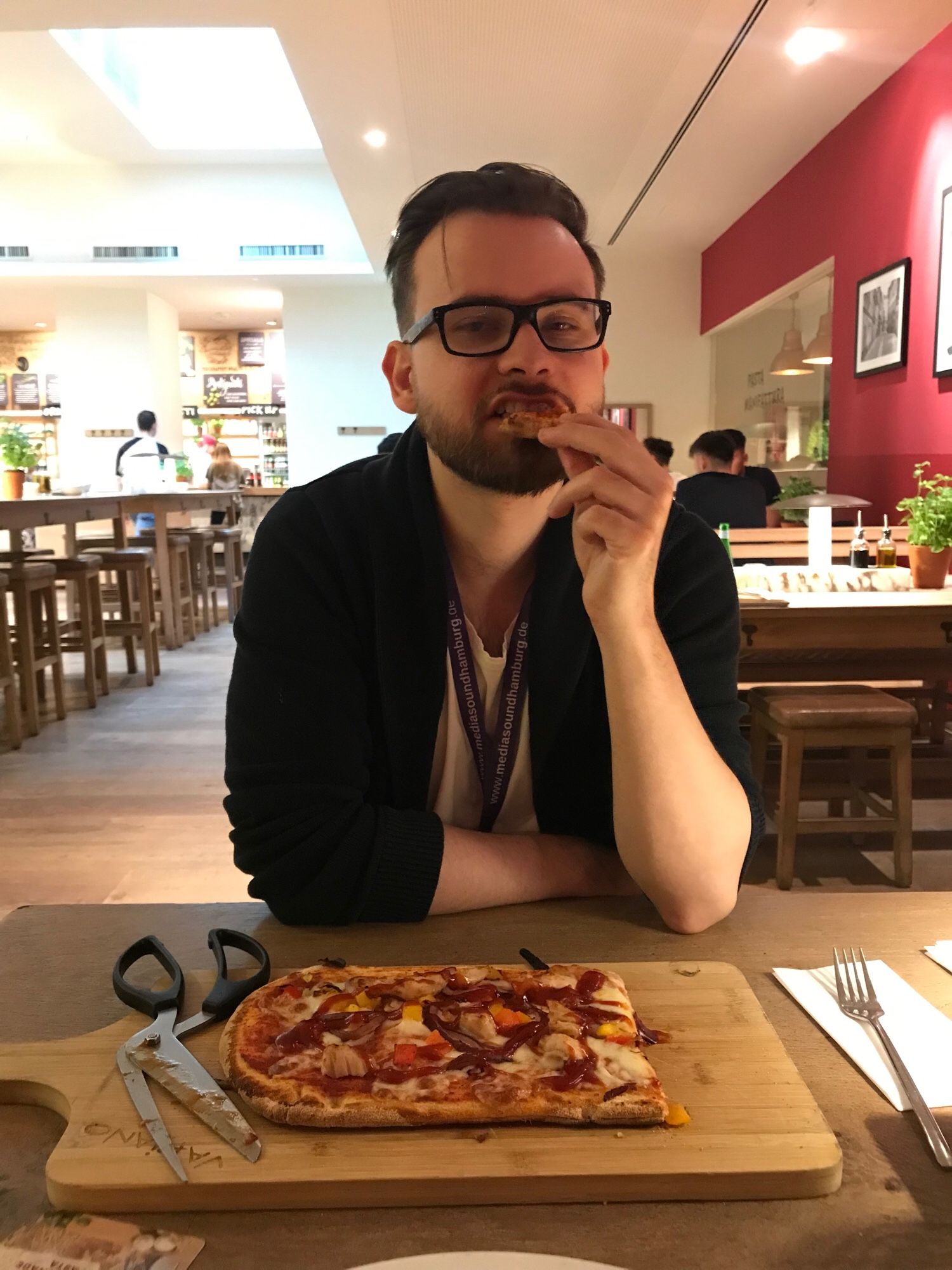 A man with dark clothes and glasses makes a silly face while eating pizza in a warm restaurant.