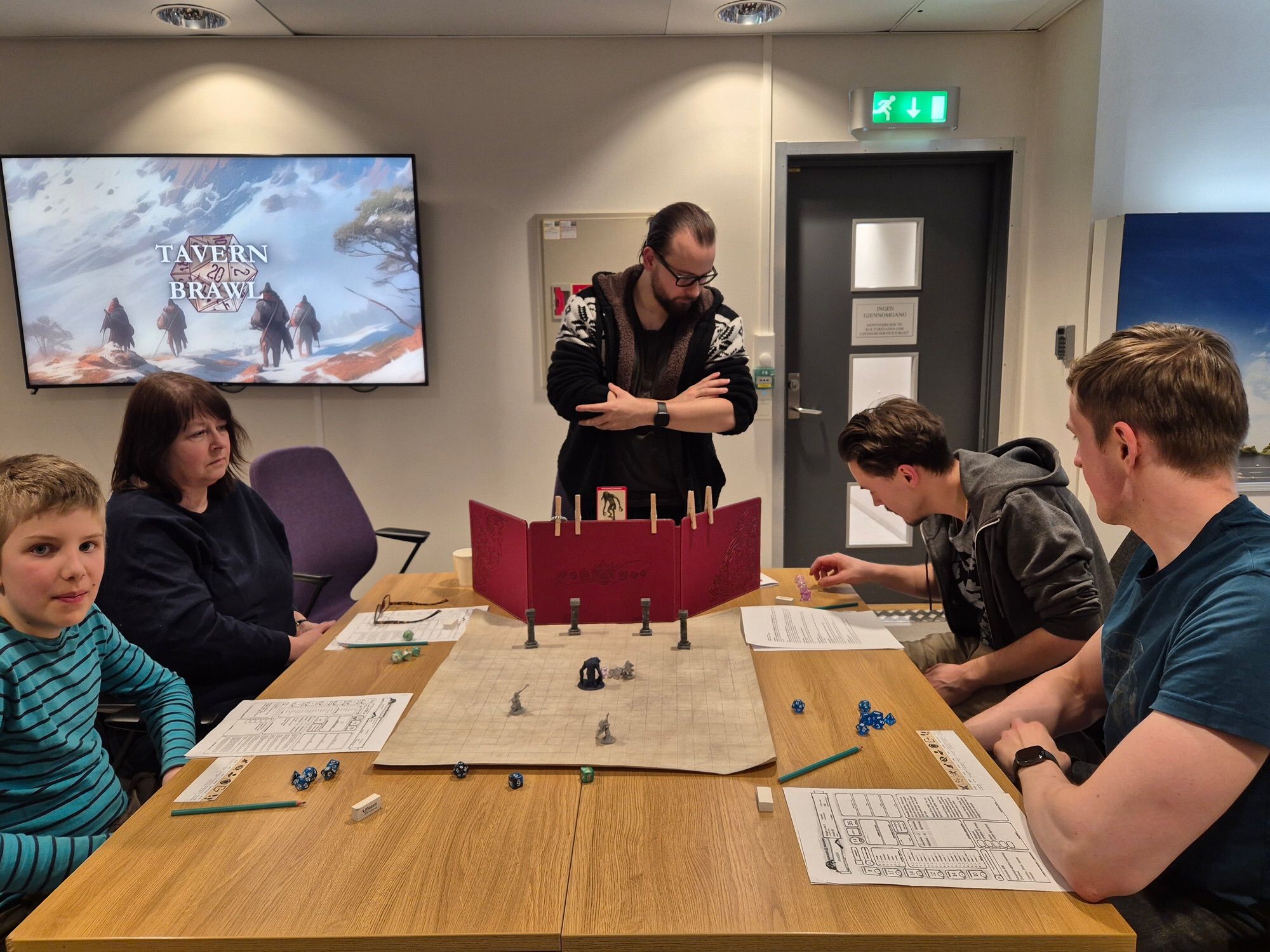 5 people are gathered around a table to play D&D. The dungeon master, standing behind a red DM screen, is looking at a die that was just thrown by one of the players.