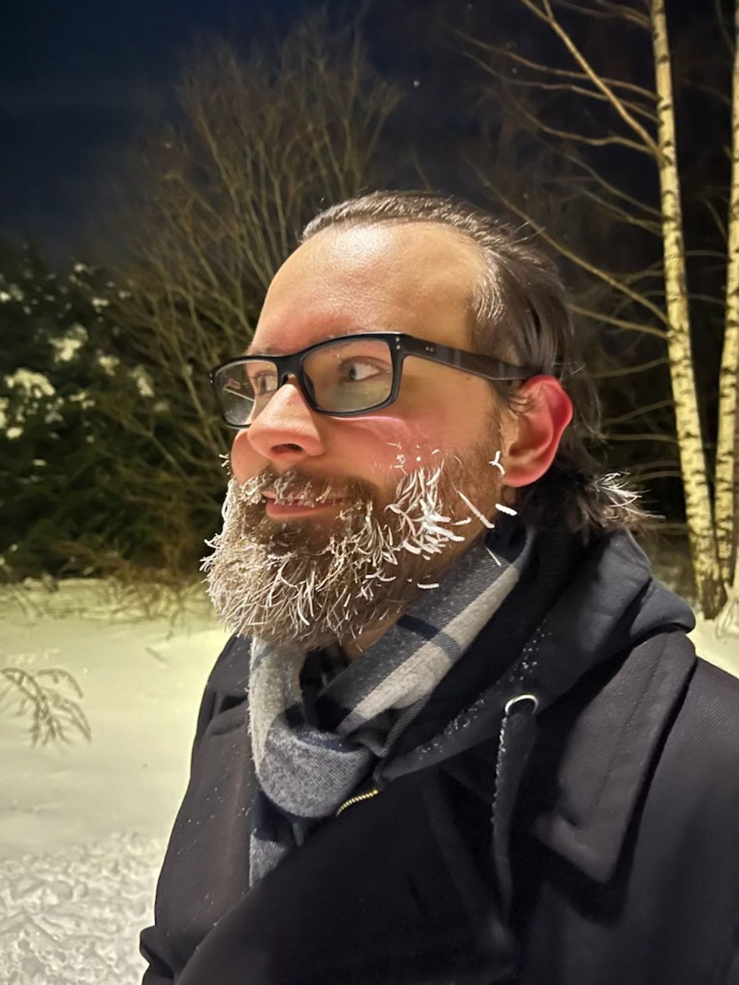 A man with dark glasses is standing in the snow, his beard white with frost as he looks off to the left.