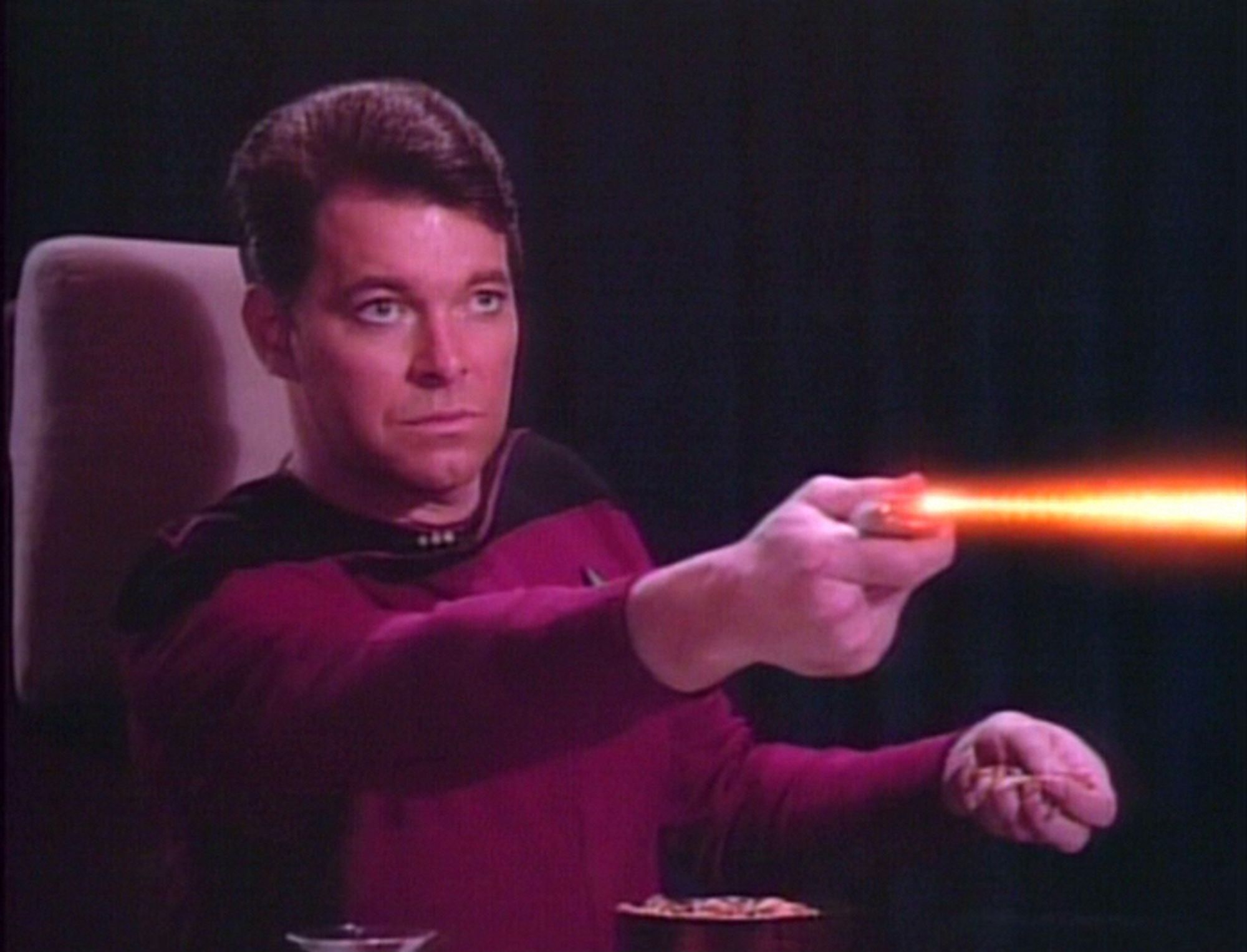 beardless riker calmly shooting a phaser toward screen right while holding space snacks in his other hand