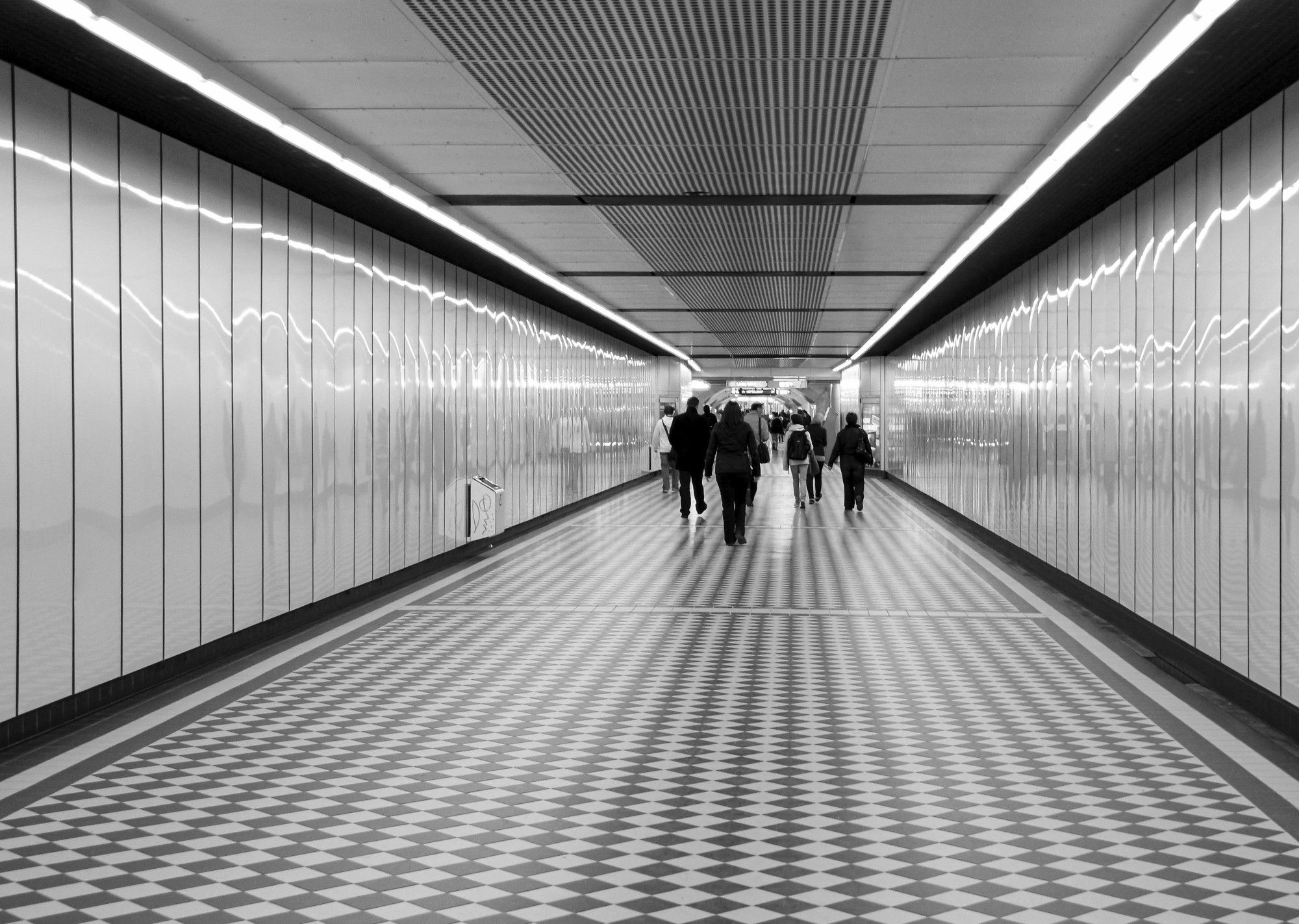 Schwarz-Weiß-Photo. Eine Unterführung, am Boden quadratische Kacheln in hell und dunkel, rechts und links glänzende helle Paneele, an der Decke außen rechts und links ein Lichtband, in der Mitte Streifen, die die Verjüngung zusätzlich unterstreichen. In weiterer Entfernung gehen Menschen.