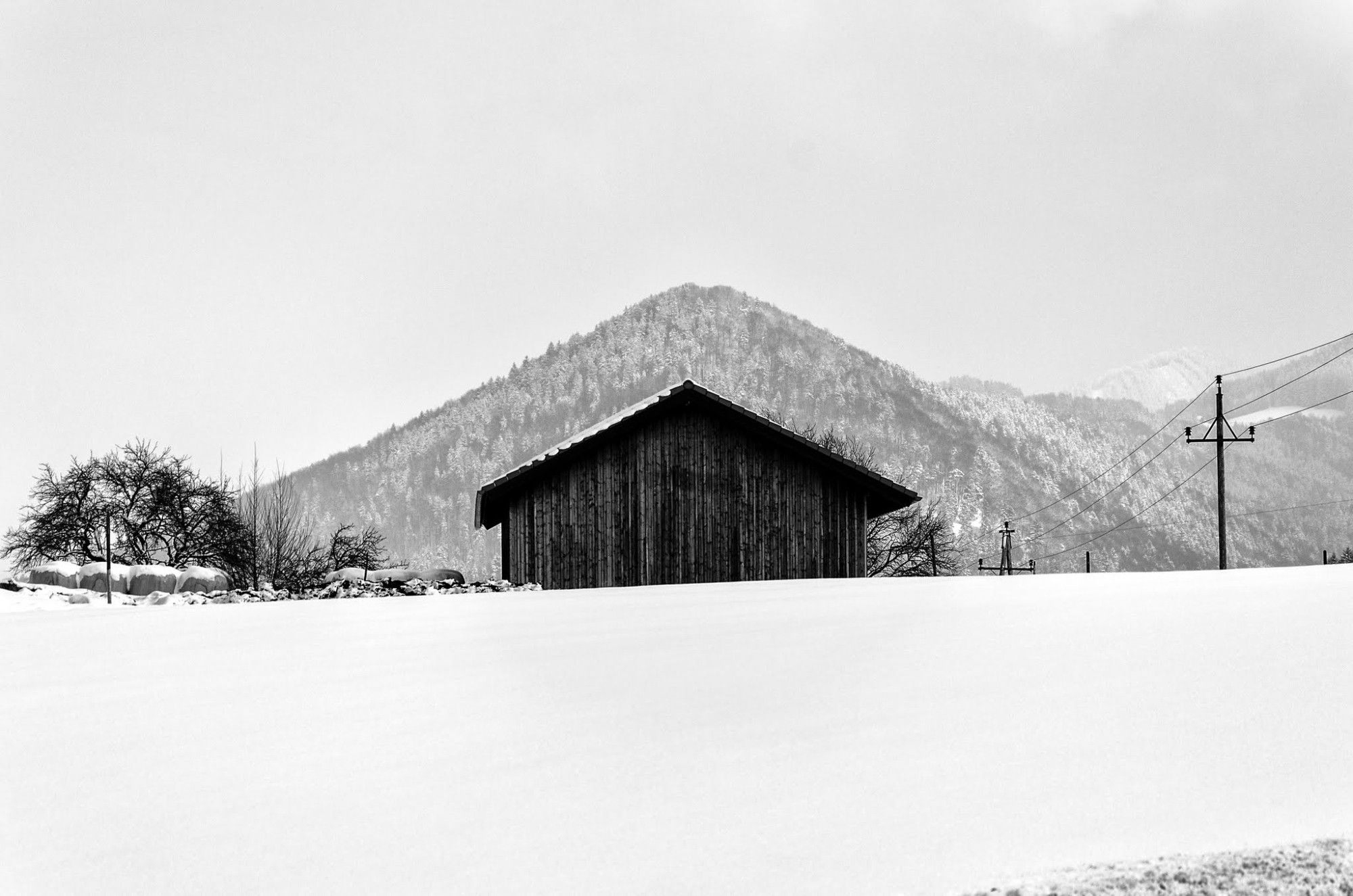 Schwarz-Weiß-Photographie. Auf einer leicht schrägen Schneefläche steht ein Holzschuppen, dessen Satteldach fast exakt den gleichen Winkel aufweist wie der Berg in Kegelform dahinter.
Schuppen und Berg sind zentral im Bild postiert, rechts davon ein Strommasten und im Hintergrund weitere Berge, links ein Baum.