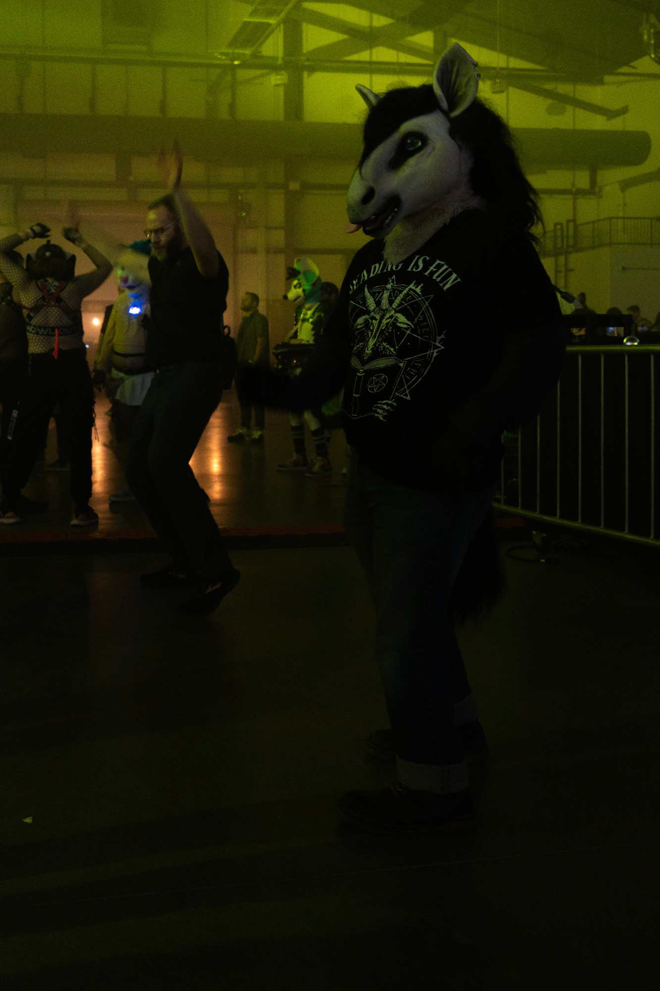 Amen and a horse costume which has a black Mane white fur and blue eyes, with a pink tongue poking out of the mouth dances in a warehouse setting. He is wearing doc martens, roll up jeans, and a t-shirt depicting a satanic goat reading a book, with the text on the shirt saying reading is fun. The warehouse has a green Halloween tent to it, with people dancing in the background, one of which is a bearded man, another which is t-shirtless person in my harness, and the person for this and back is a white and blue candid. There is also the portable metal barrier for the Rave effect steam Behind The Horseman