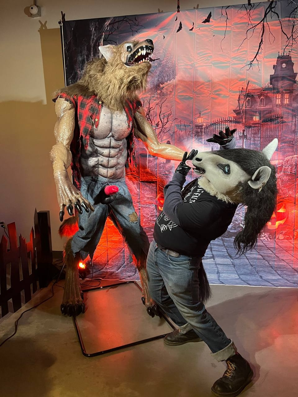 A man in a horse costume comedically recoils from a muscly werewolf mannequin which has a red dog boner poking through its pants. The horse costume has a black man, wait for in blue eyes, and a pink tongue pokes out of the mouth. He's wearing a black T-shirt with a cult symbols on it, blue jeans with the hems rolled up, and Doc Martin boots with rainbow laces. The werewolf wears a tattered red and black denim best and shredded blue pants with no shoes. Behind both figures is a vinyl backdrop of a haunted house, complete with glowing jack o lanterns on the front