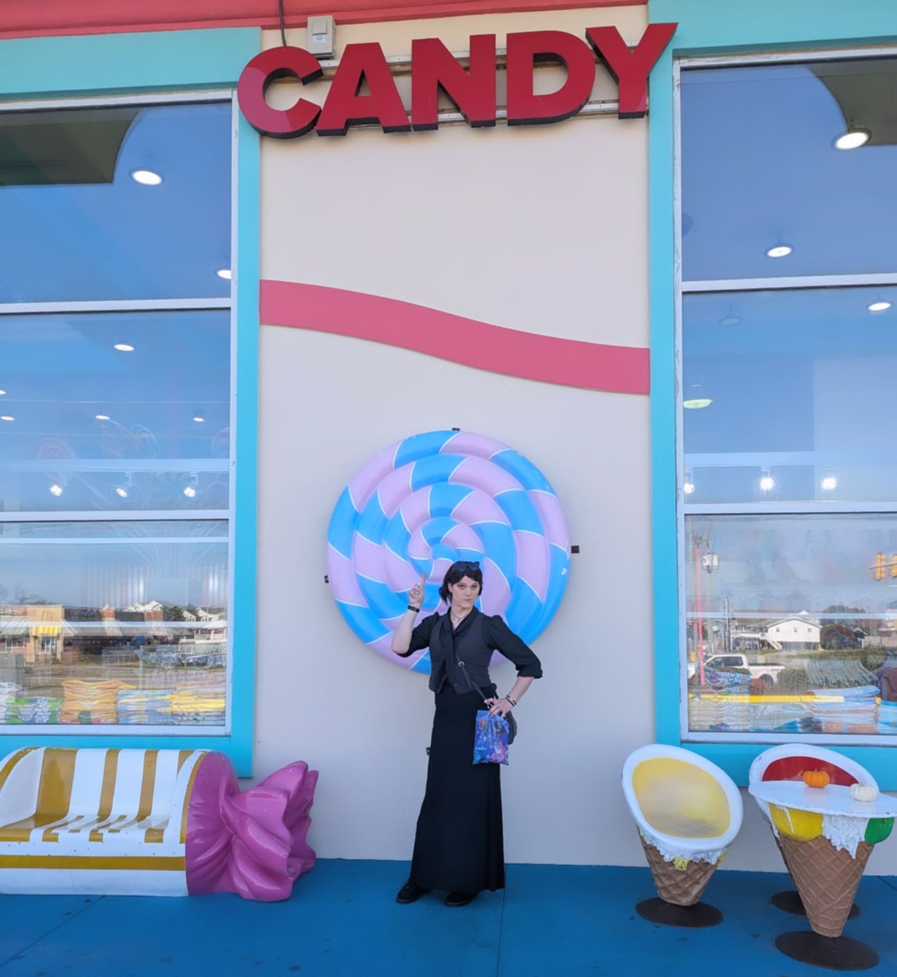 A photo of me, Velcro Candy, standing in front of a large, red neon sign reading "CANDY."
The sign is not currently lit. 
I am wearing a black blouse, gray vest, long black vest, a gray headband, black corduroy Doc Martens, black, heart shaped sunglasses on my head, a digital watch, a bracelet, red eyeshadow, pink press on nails, black eyeliner, false eyelashes, two rings, and three earrings in each ear. I have a purse over my shoulder and I'm clutching a shopping bag with my left hand which is also resting on my hip. My right hand is raised with my left index finger pointing upwards, towards the aforementioned neon sign.
Around me are confection shaped decorative items of furniture. 
I have brown eyes and a pale complexion. My hair is black. I'm actually older than I look/admit to being. The ground is beneath me, as of to indicate I'm not in zero gravity. There are no birds of prey visible in the photo.