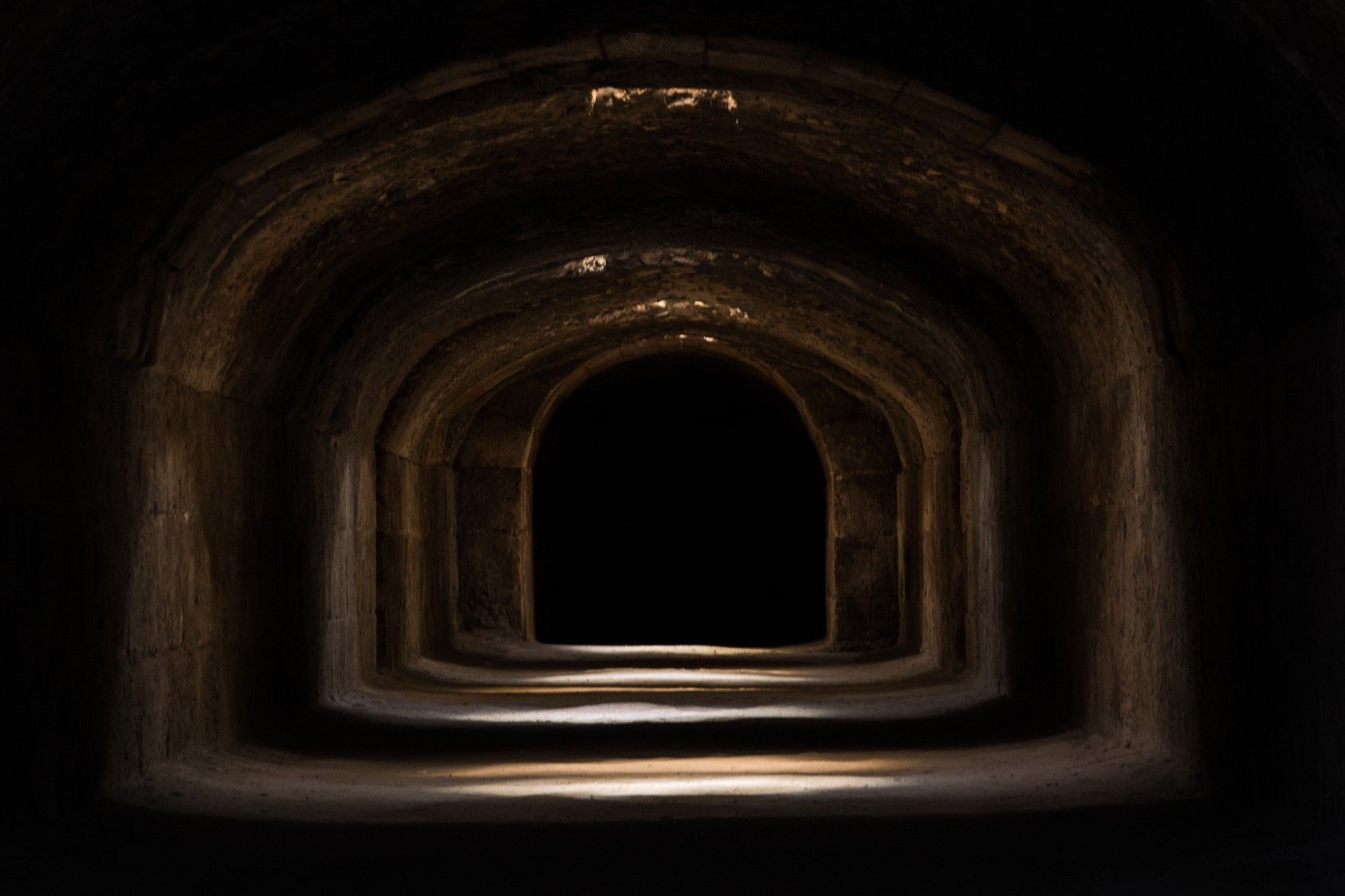 Picture of a broad, underground tunnel with deep shadows between the four pools of light from unseen ceiling skylights that lead down the tunnel. The walls are large, blocks of sandstone and you can see the stone arches going down the tunnel, leading to what is probably a wide arched doorway, but it is lost in the shadows.