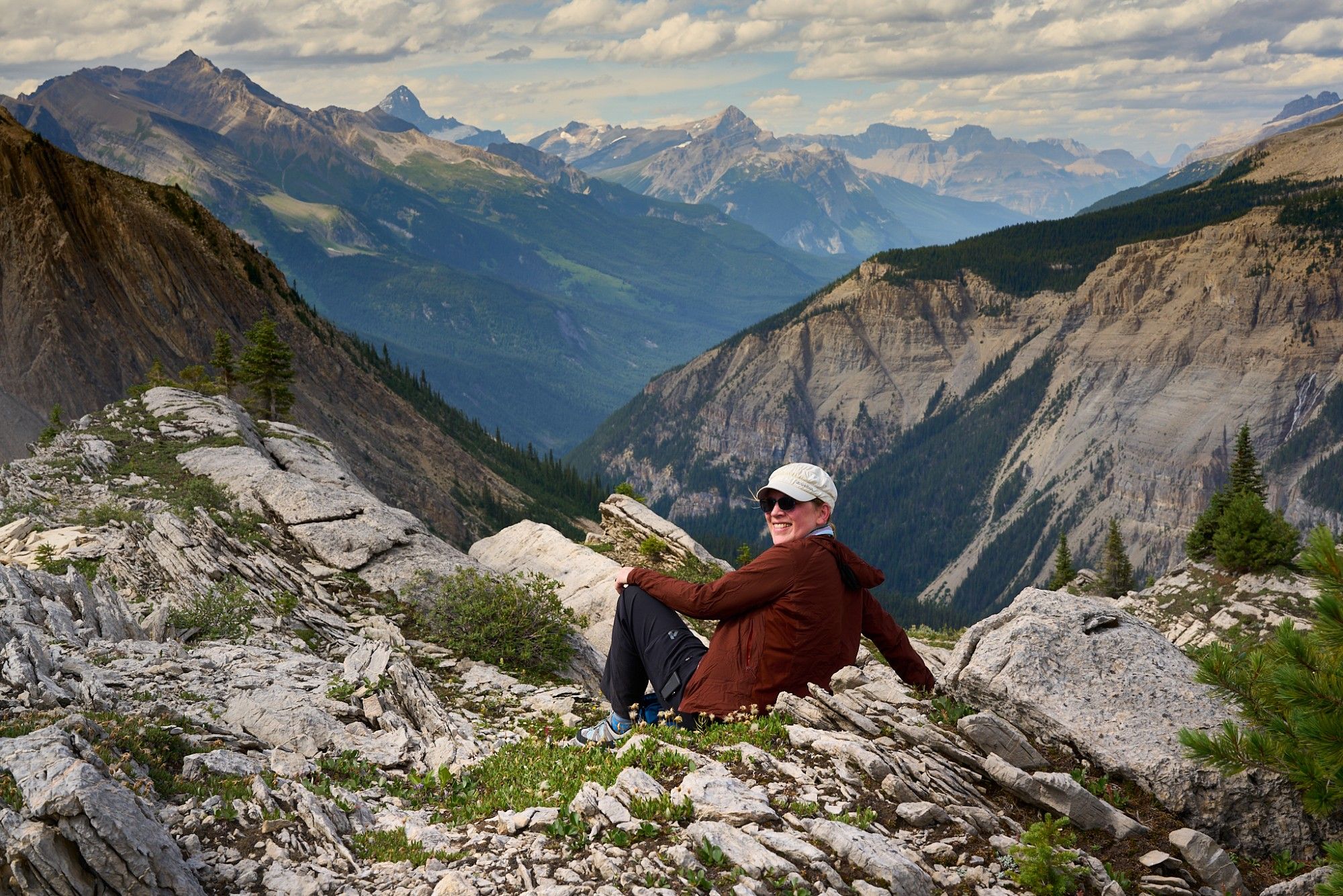 View from the ridge at the west of the bowl near Mistaya Lodge. LMC sits in a rubble field, in the distance is a valley and ridge of mountains. Clouds fill the sky.