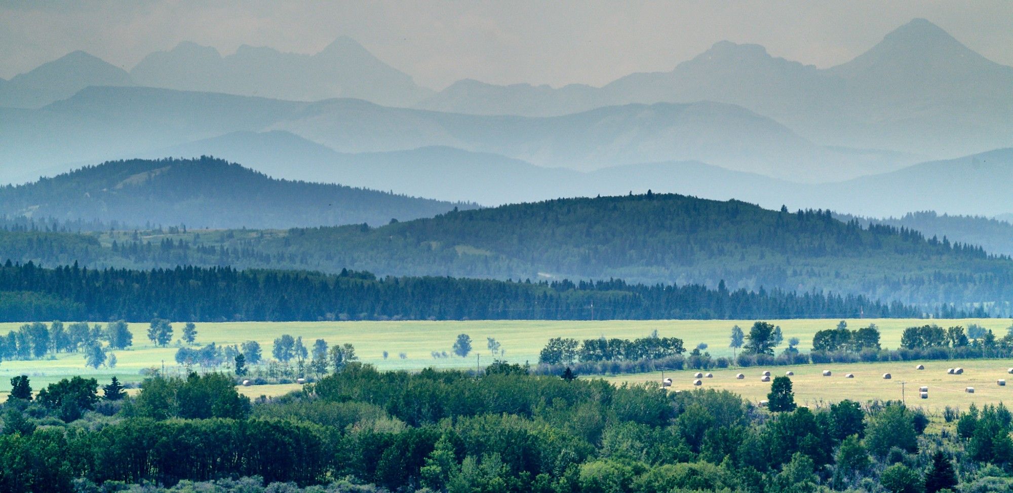 Smoke fills the Alberta foothills. A series of layers is visible stretching to the horizon. Each one is progressively more obscurred than the previous. Smoke seems to cling to the valleys in between.