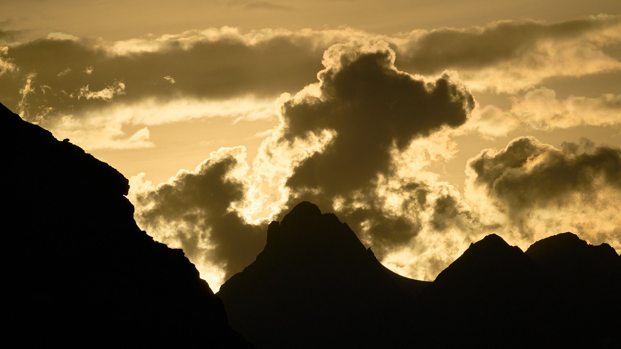 Sunset looking west from the Mistaya Lodge helipad. A ridge of mountains stretches from left to right and are silhouetted by the Sun that has set behind them. Clouds rise up in the gold of the sky.