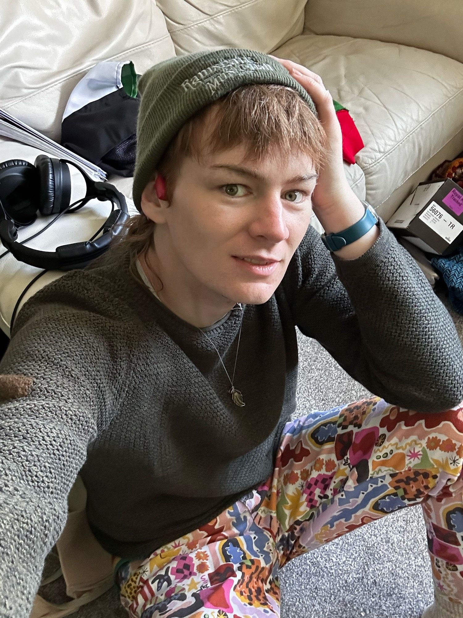 Reverie, sat on their floor in multicolour jeans and a green jumper and beanie. Feather necklace. Blue watch strap visible. Like if a boy was a girl.