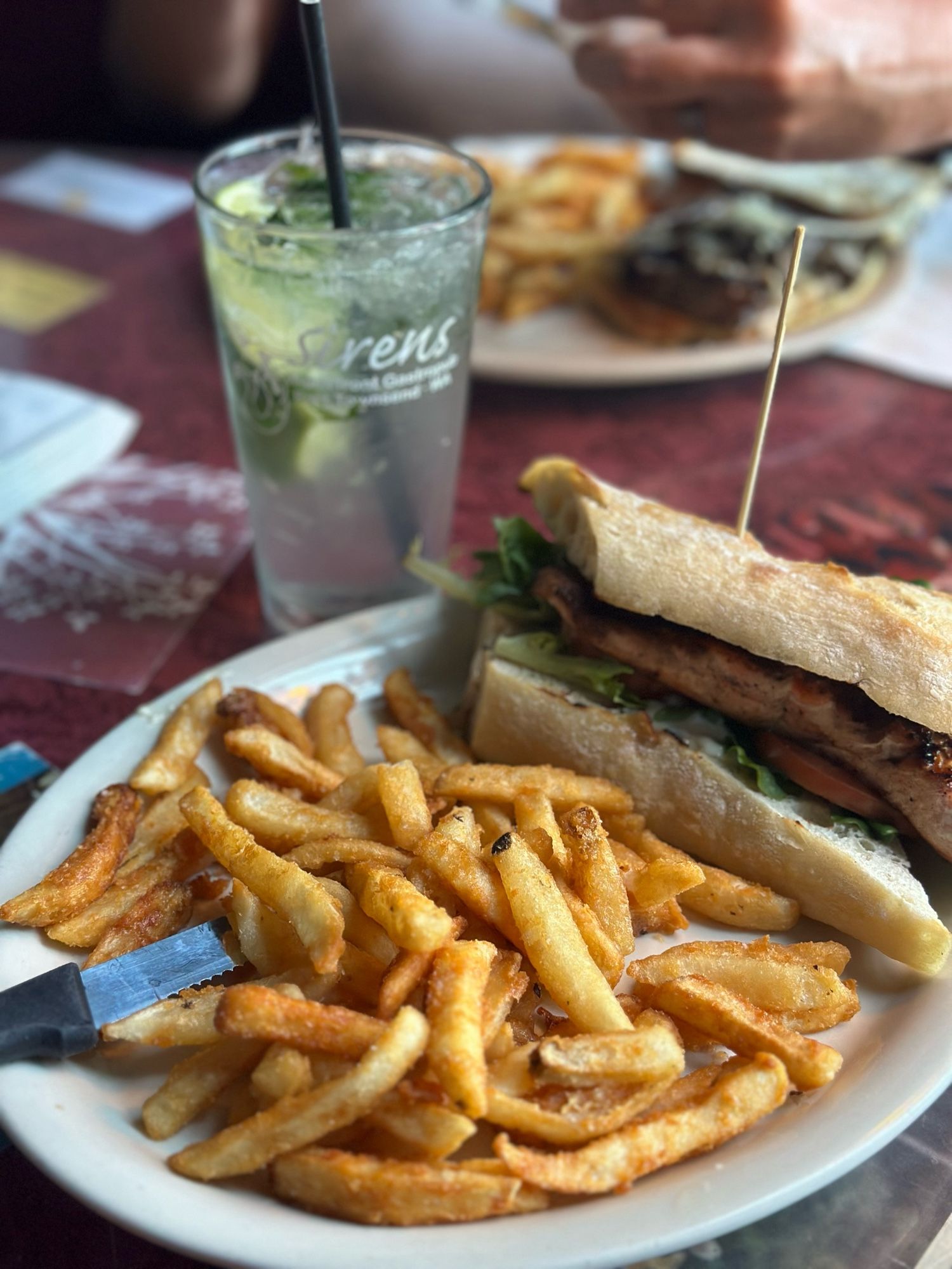 A photo of a plate of fries and a grilled salmon sandwich next to a mojito. The drink glass says Sirens on it