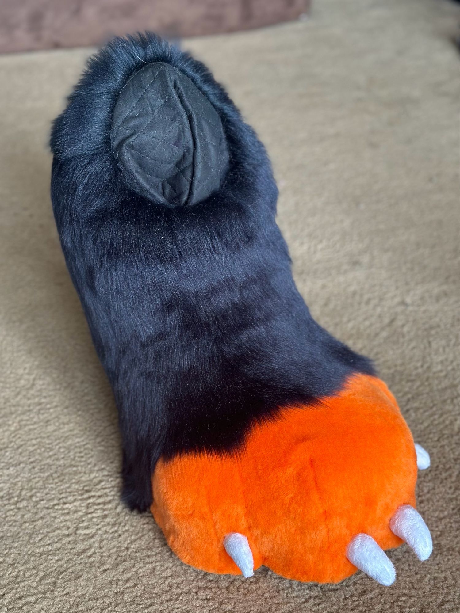 A photo of a pair of digitigrade fursuit feet. The toes of the foot are orange with small white claws and the foot/calf of them is black