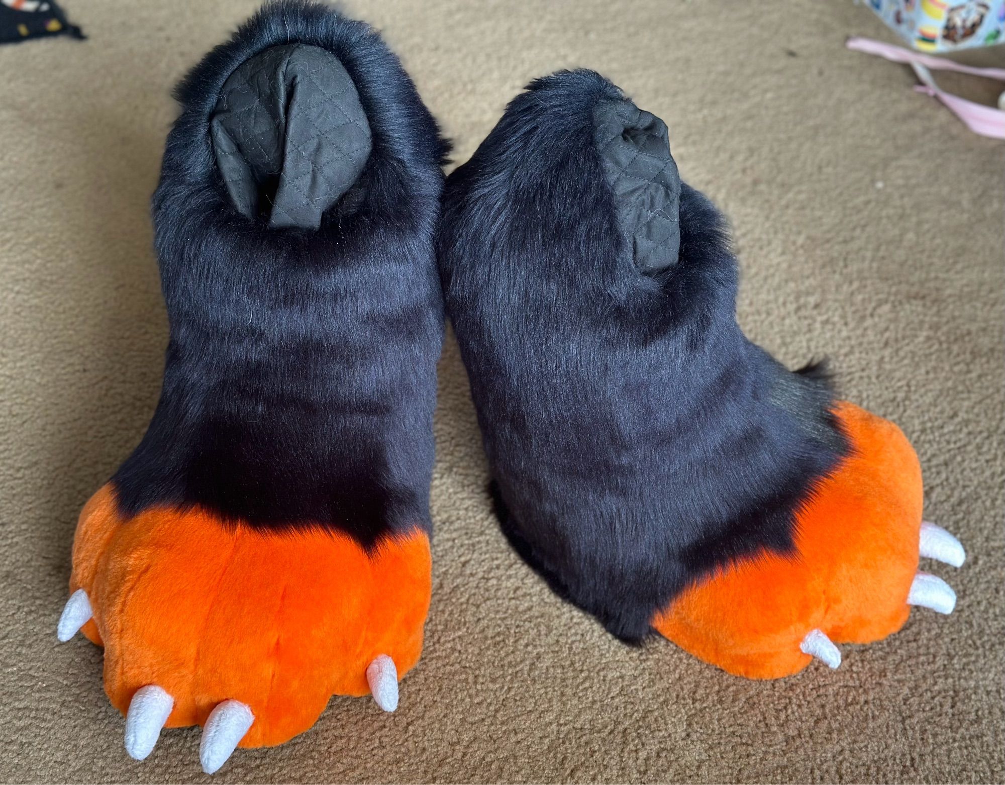 A photo of a pair of digitigrade fursuit feet. The toes of the foot are orange with small white claws and the foot/calf of them is black.