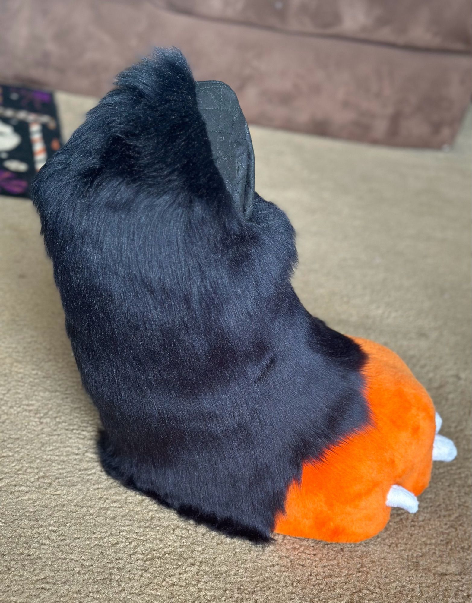 A photo of a pair of digitigrade fursuit feet. The toes of the foot are orange with small white claws and the foot/calf of them is black