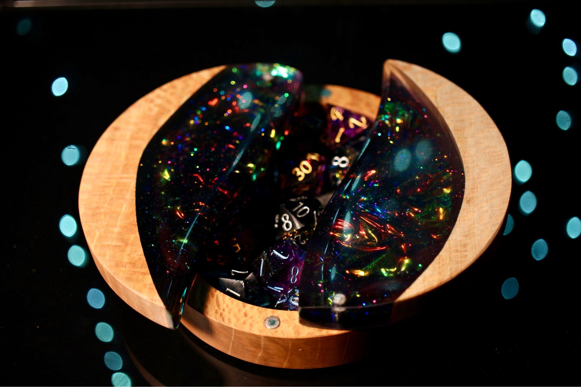 Round Sycamore dice box with a large oval cut out, filled with iridescent foil of yellow, green, orange and blue as well as dark epoxy. Shown open with the two halves of the circle that comprises the top split down the middle revealing dice inside.