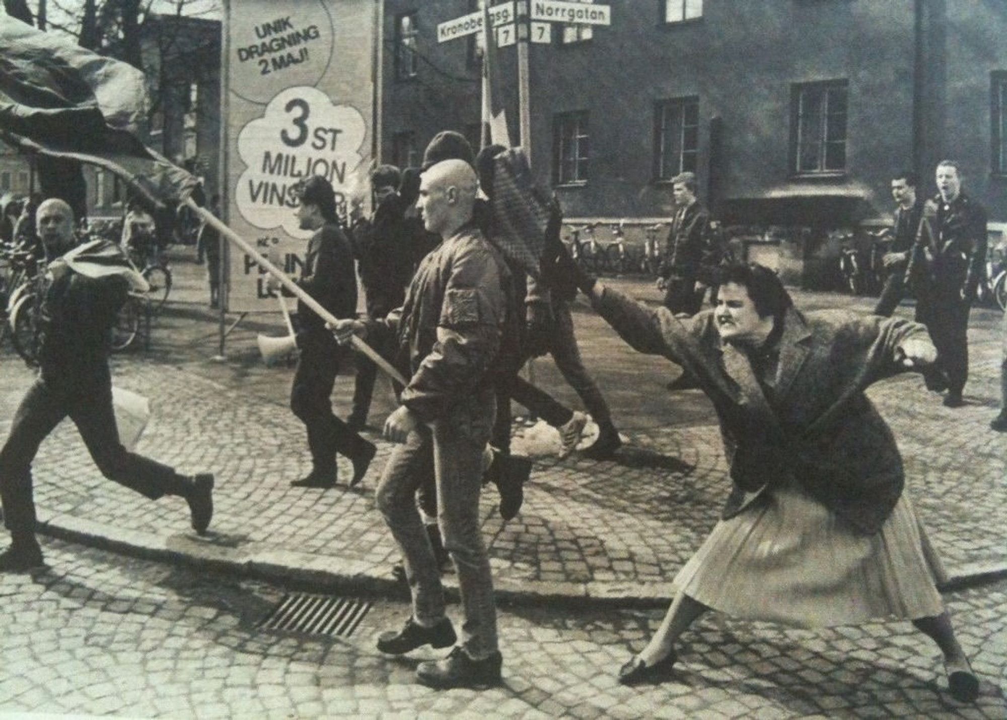 Schwarz-Weiß Foto: Ein großer Platz mit Kopfsteinpflaster. Eine Frau in einem weißen Rock und dunkler Jacke, sowie dunklen Haaren steht breitbeinig, mit wütendem Gesichtsausdruck hinter einem Neonazi, mit Bomberjacke, Jeans sowie Glatze, und schlägt ihn mit ihrer Handtasche in der rechten Hand auf den Hinterkopf. Im Hintergrund sind mehrere Personen zu sehen, die die Situation teilweise beobachten und zum Teil den Neonazis angehören