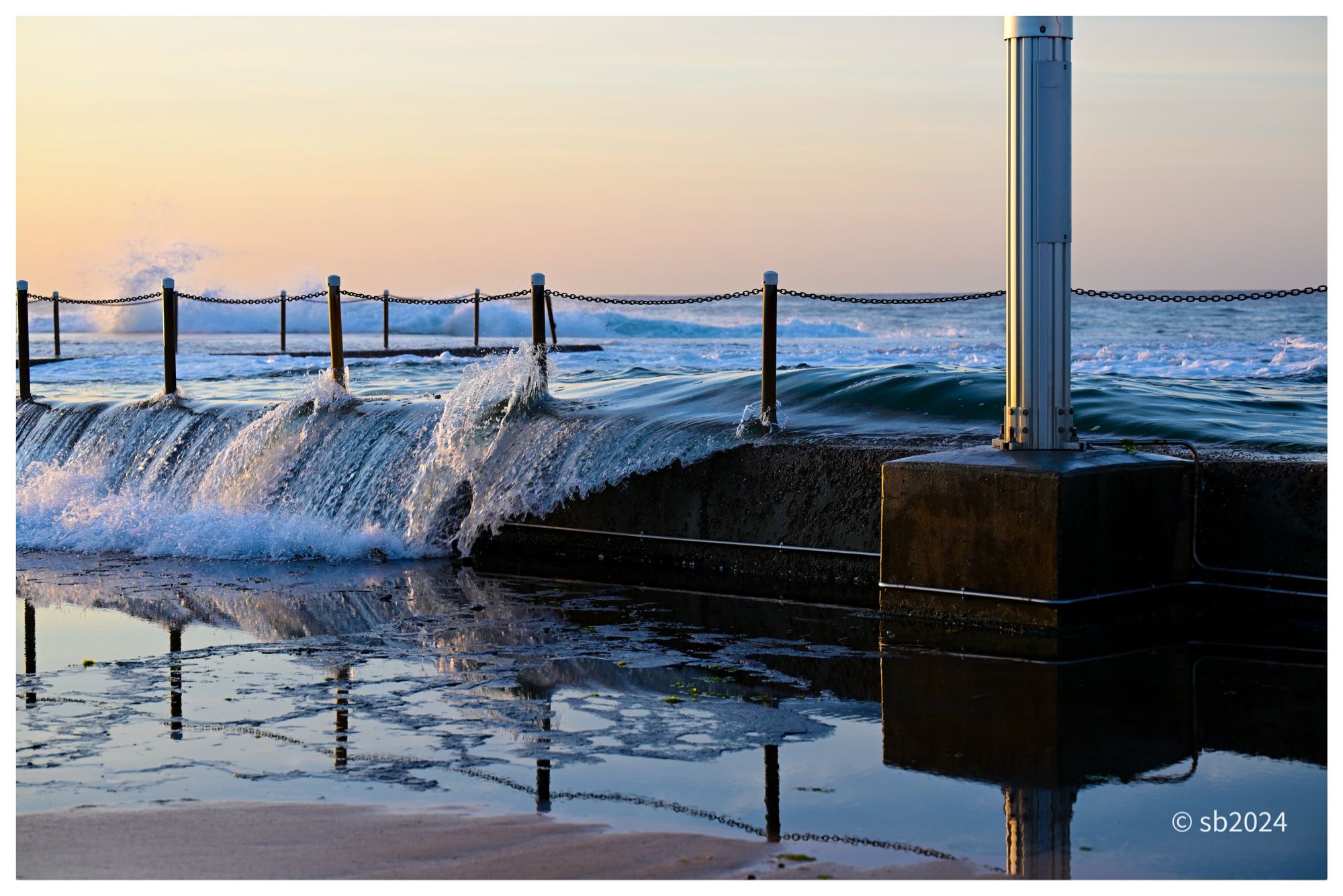 In the foreground is a mirror reflection of the edge of a rockpool, its fencing,  and the base of a tall silver light on the right. Waves surge over the rockpool. A clear morning sky in the background.