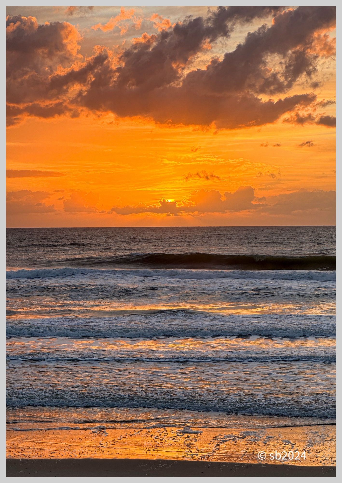 Reflection of a golden sunrise on the wet sand. Waves roll to shore. The sun rises over the horizon partially obscured by a long cloud. Large grey clouds drift above the sun.