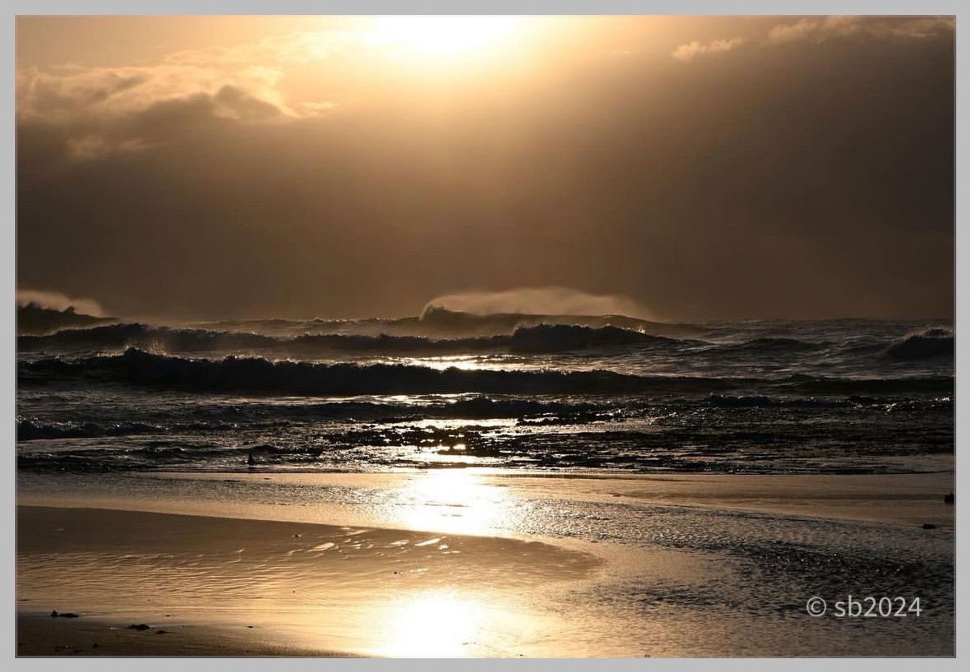 The sun rises over the sea partially obscured by clouds creating orange drifting across the horizon. Large, long line of waves crash to shore. The golden hues of the sun are reflected in the wet sand in the foreground.