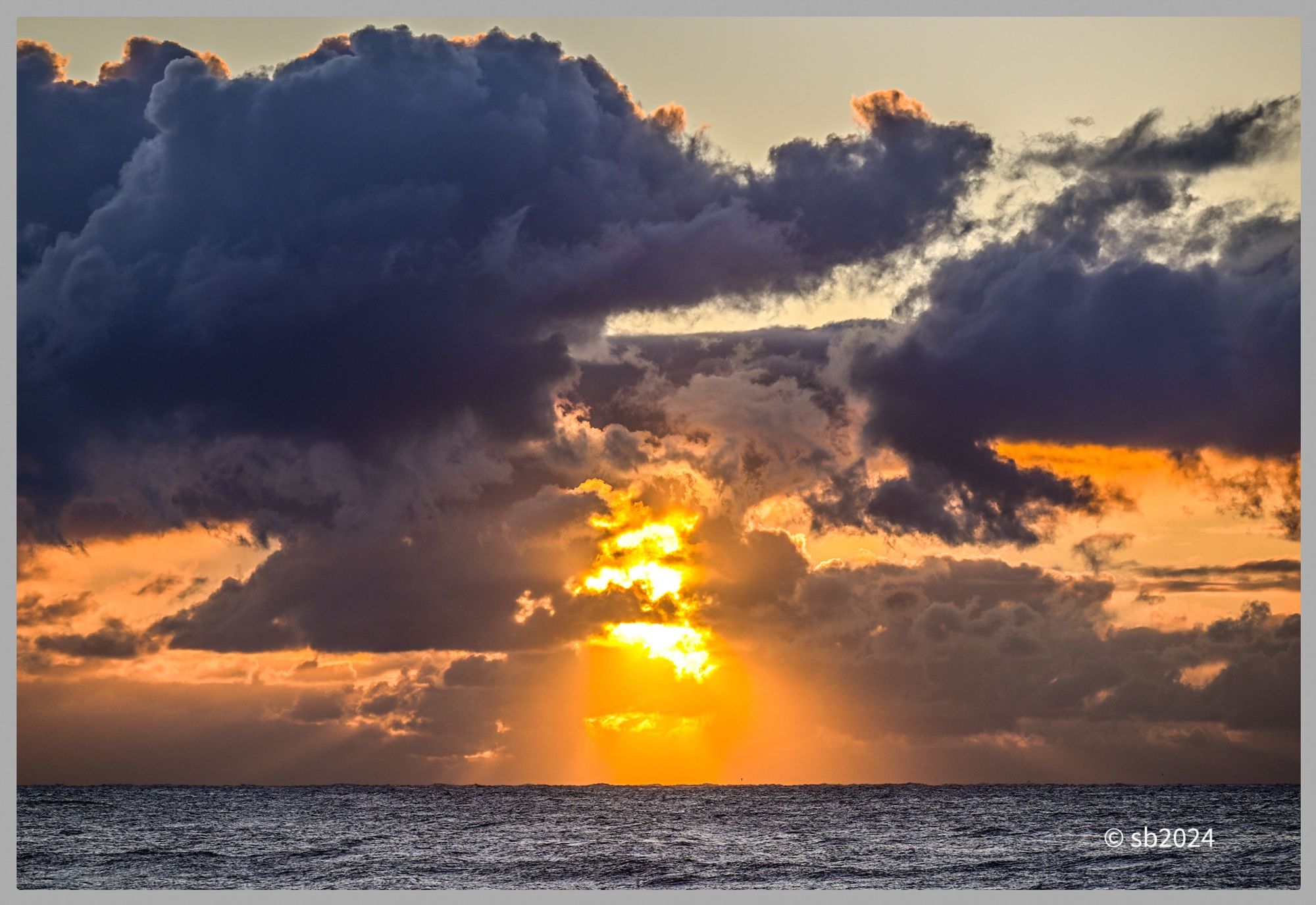 Foreground shows the sea, the sun rises over the horizon partially obscured by clouds. Rays of sunlight stream through breaks in the clouds creating orange yellow glow.