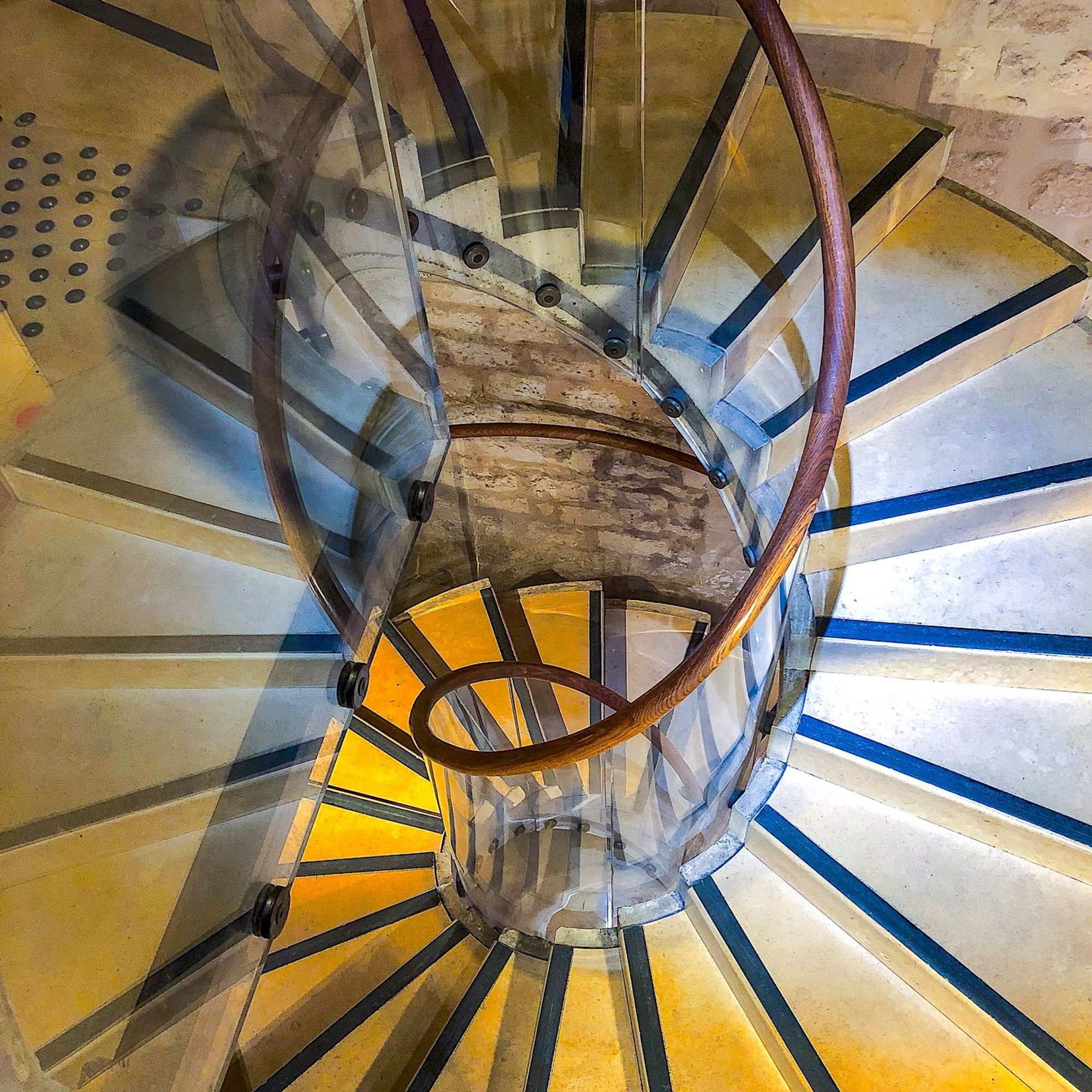 A spiral staircase with stone steps a wooden handrail and clear glass safety panels joining the two. The stairs lead downwards to the basement exhibition room of "Mundolingua" the Museum of Languages in Paris.