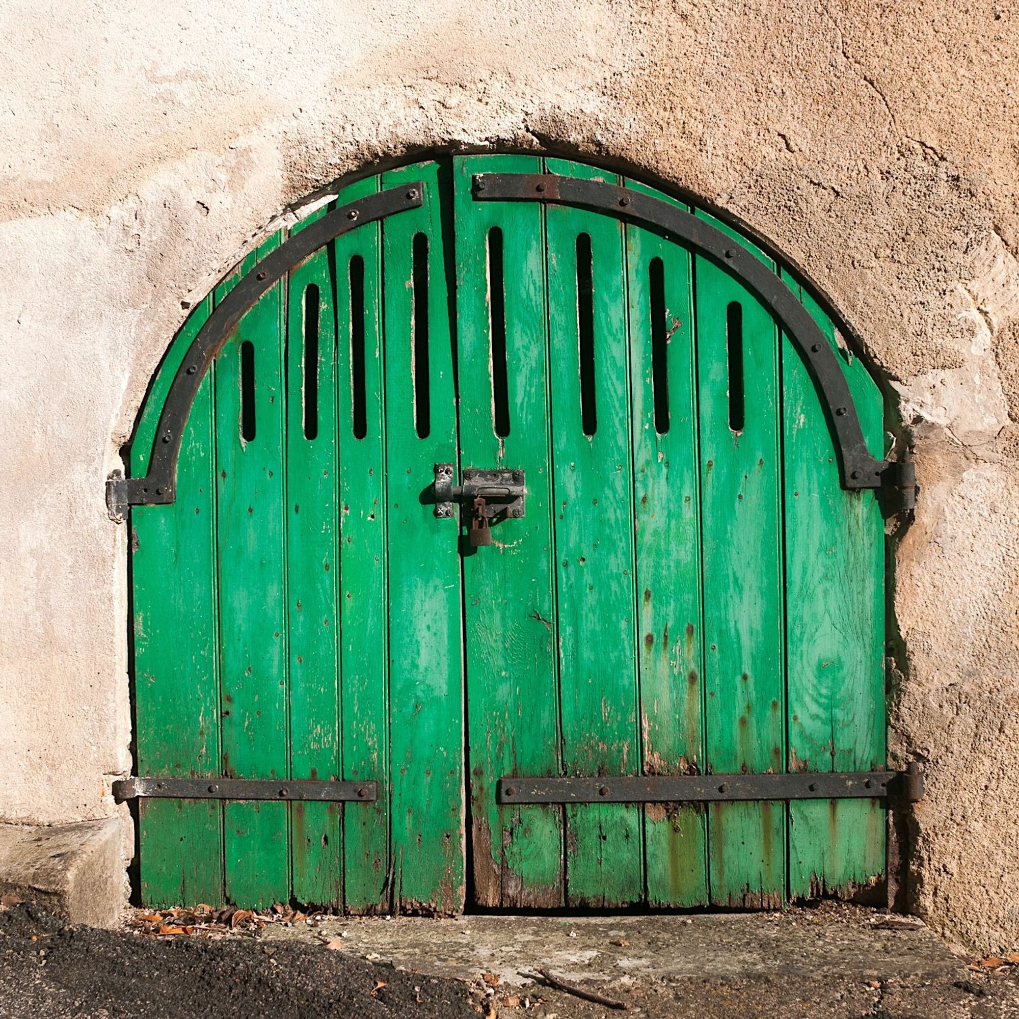 Double doors painted bright green curved at the top to fit an arch in the stonework. Large hinges, curved at the top to follow the flow of the arch, and vertical slots in the doors for air circulation.