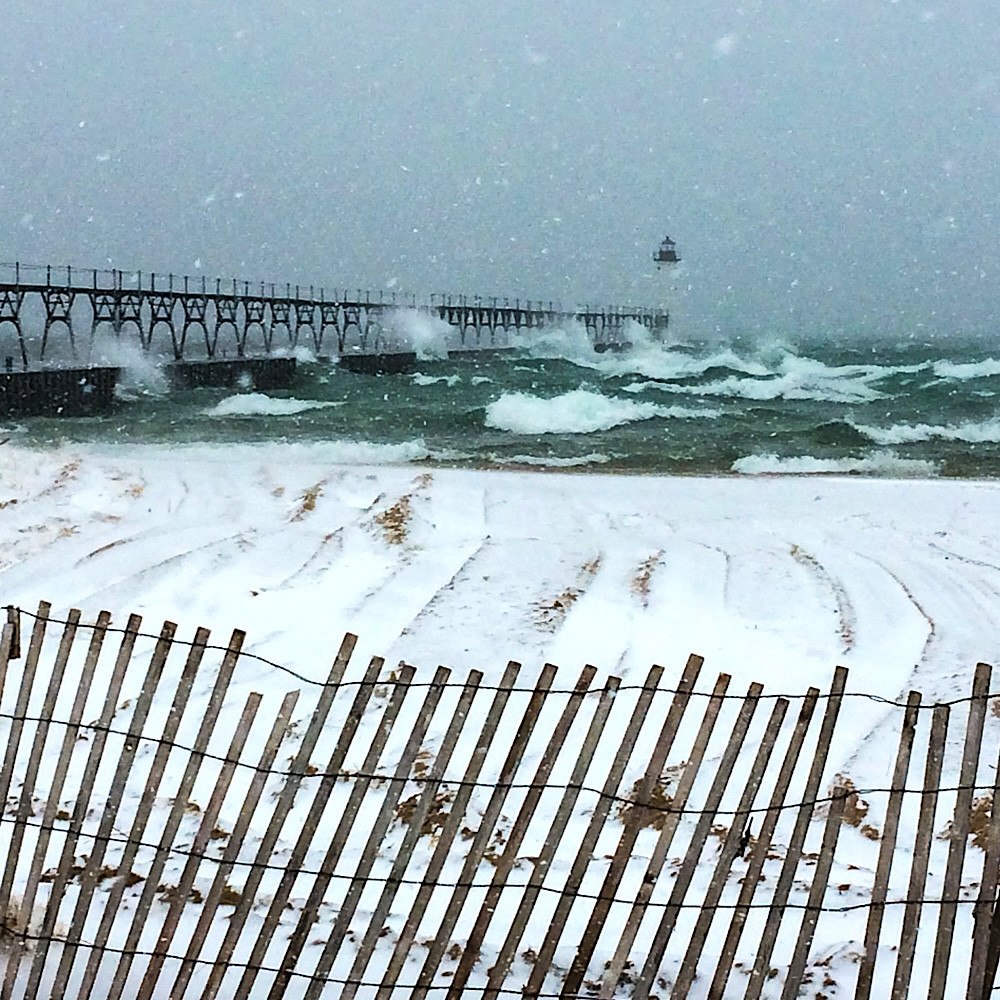 The shoreline at Manistee North Pier Lighthouse on Lake Michigan in the USA taken in mid-April during a freezing snowstorm. The waves are cresting with white foam against the pier and lighthouse; the beach is covered in snow and the wooden paling fence in the foreground is leaning in the gale-force wind.