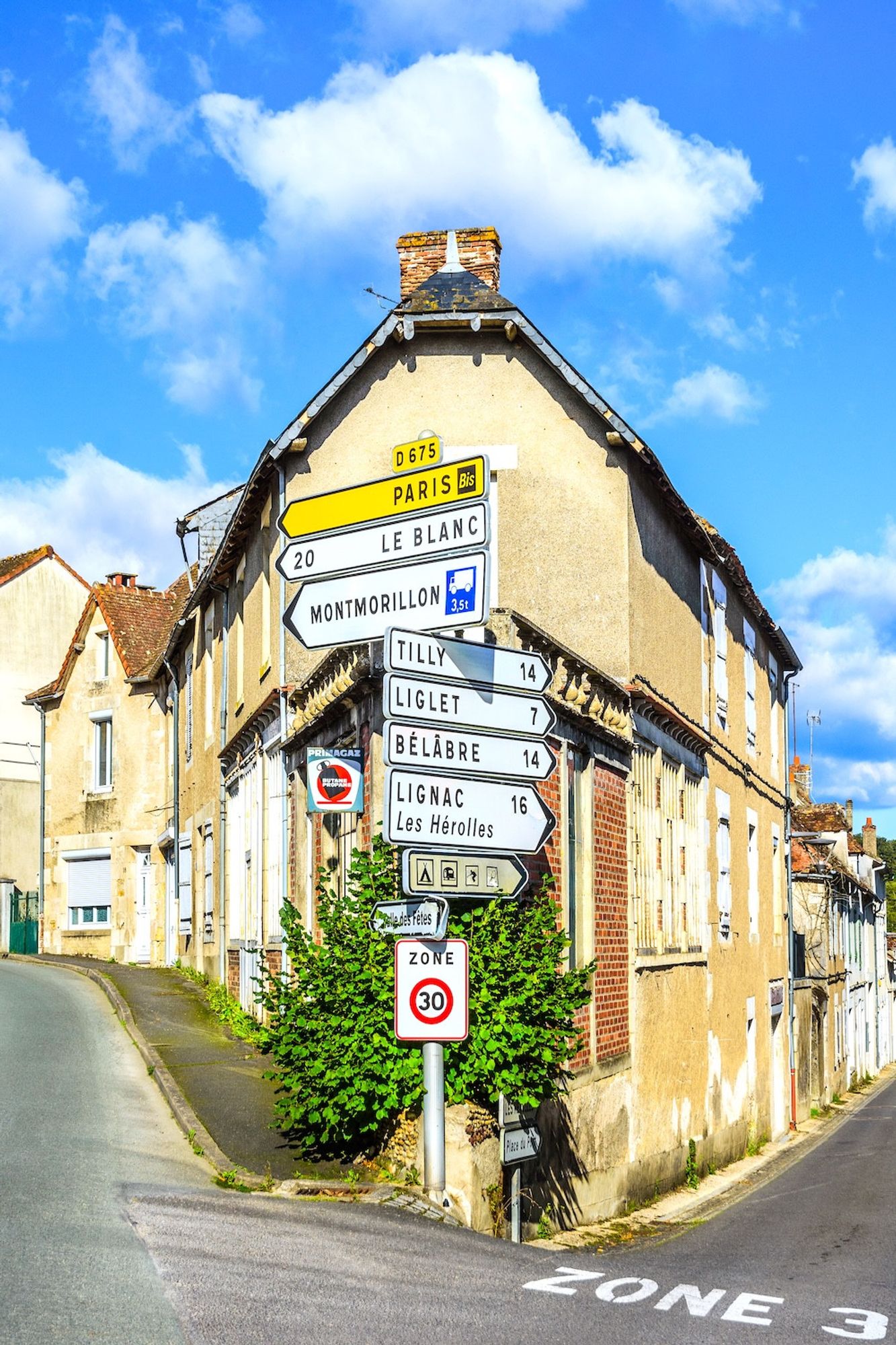 Multiple directional road signs on a post in front of a wedge-shaped corner house at a road junction in the small town of La Trémouille in the Vienne departement of central France. The road junction is on a hill in a 30 kilometer per hour restricted area of the town. Photo taken on a sunny day with a blue sky and fluffy white clouds.