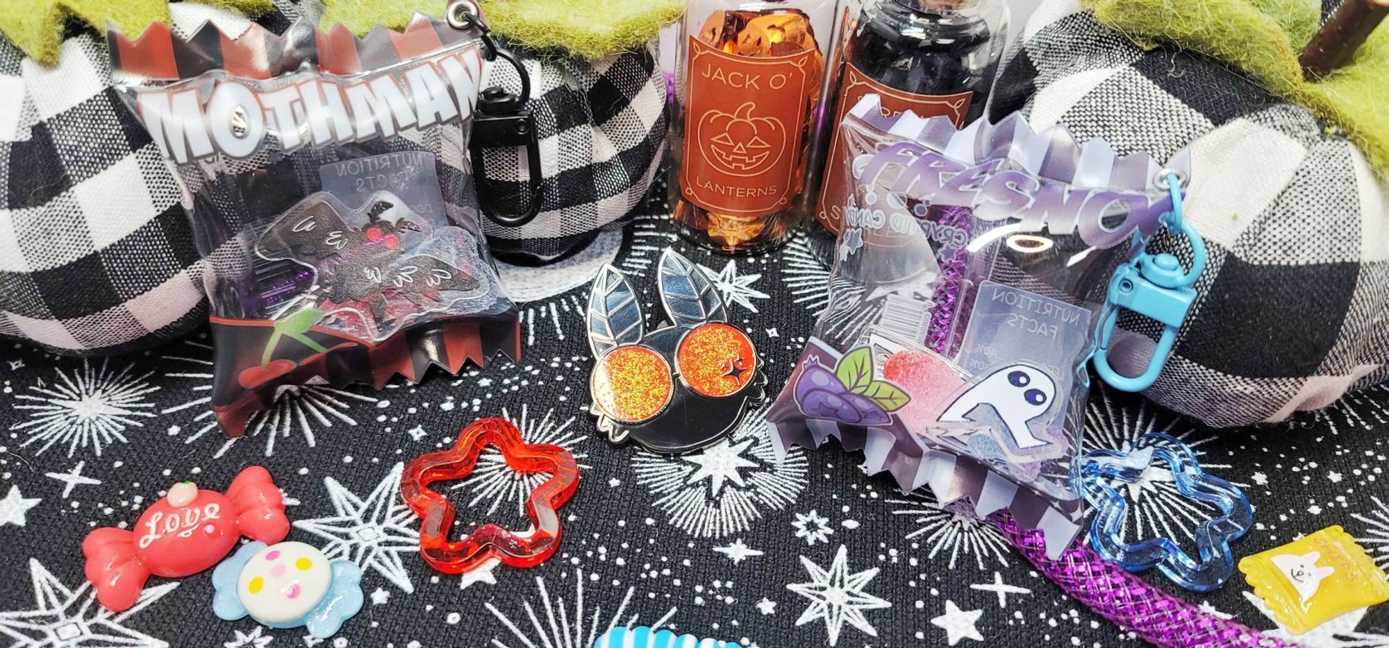 A cute little halloween spread containing some stuffed pumpkins, little resin candies and stars, as well as some bottles containing glitter. There are 2 candybag charms -- one mothman and one fresno nightcrawler themed, and a mothmanpin in the very center.