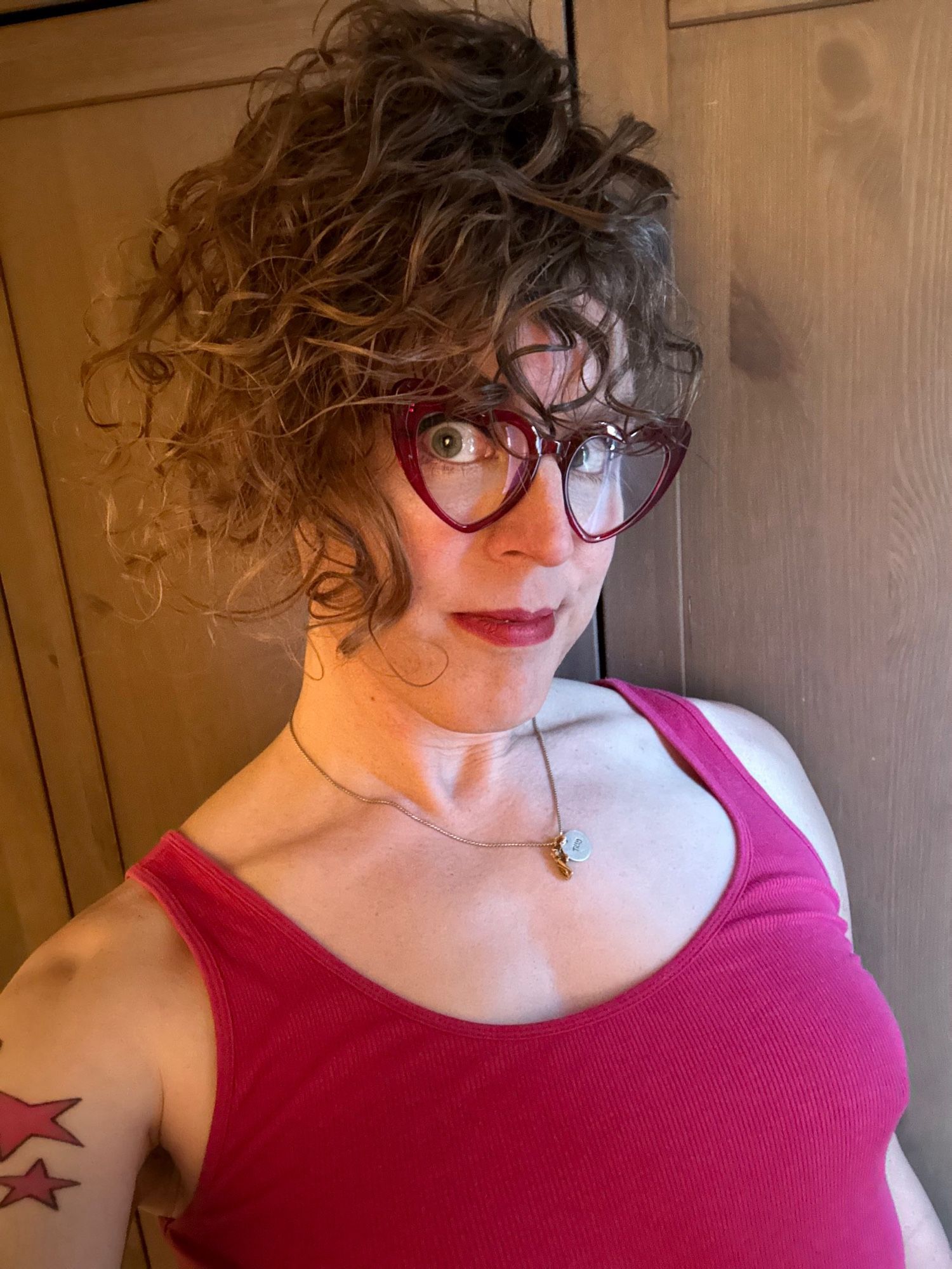 me in a pink pajama tank and red heart shaped glasses, with my hair up in a pineapple that somehow looks very windswept, despite the fact this was taken indoors, because the curls do whatever the hell they want and I can’t stop them